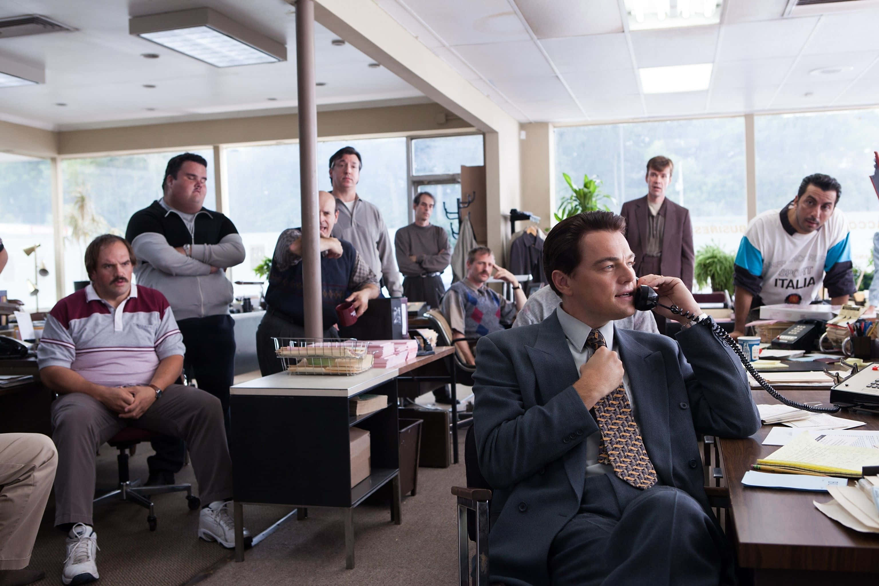 Wolf Of Wall Street 桌布 壁紙 — 李奧納多·狄卡皮歐坐在雜亂的辦公桌前打電話，周圍有一群男人專注地看著他。