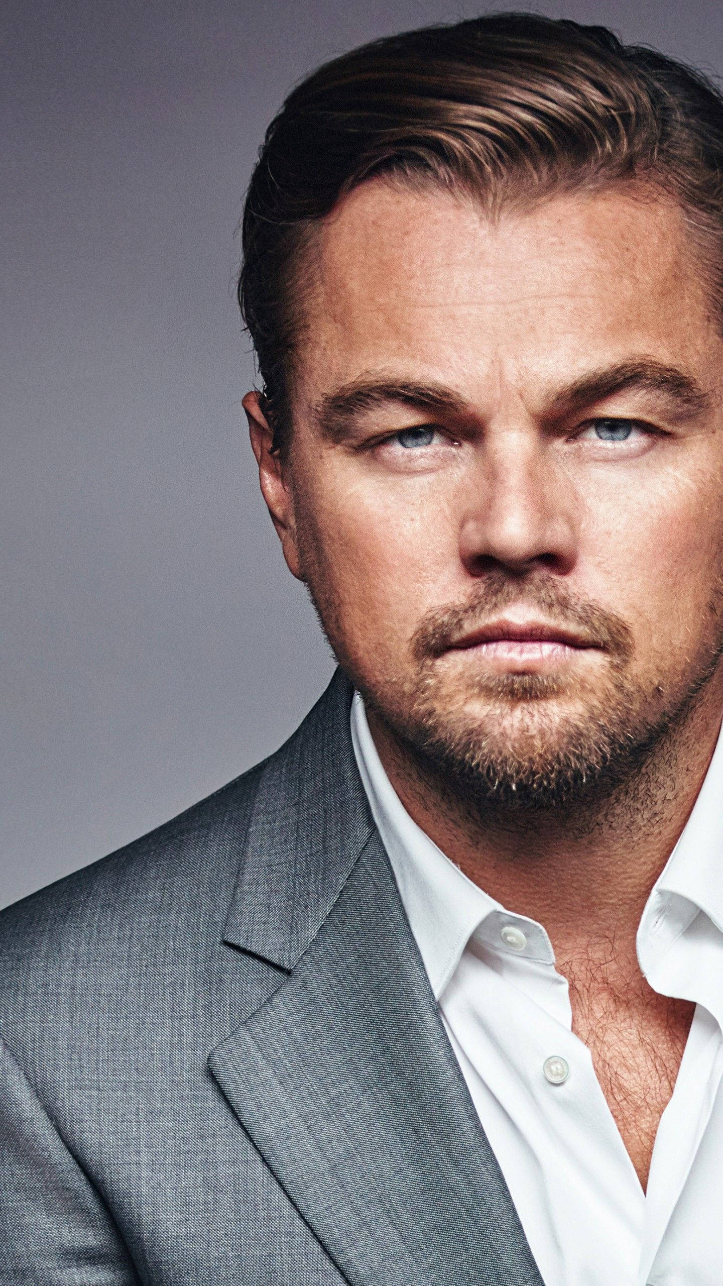 Dicaprio 桌布 壁紙 — 李奧納多·狄卡皮歐的close-up肖像，擁有強烈的藍色眼睛，穿著灰色西裝和白色襯衫，背景為中性灰色。