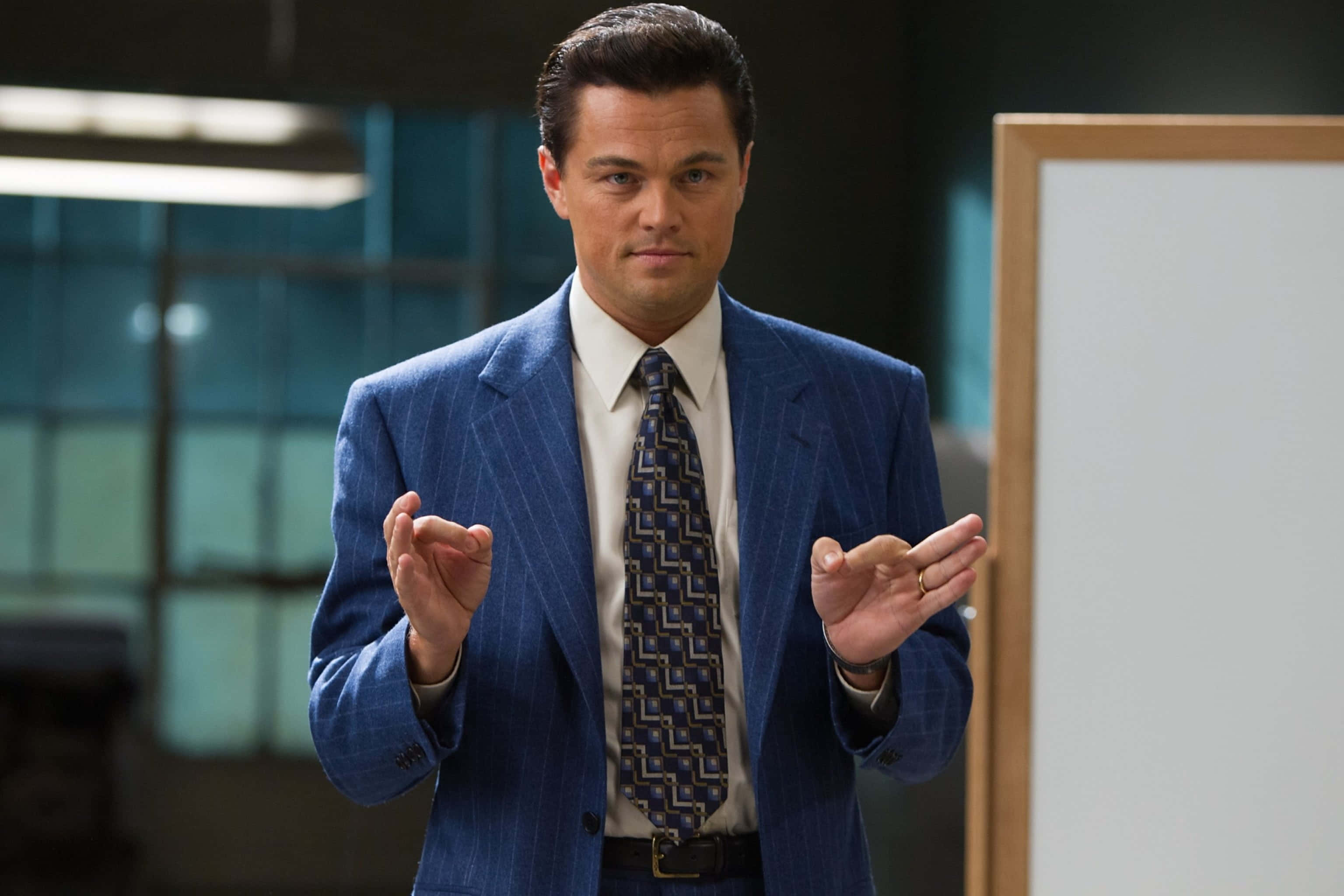 Wolf Of Wall Street 桌布 壁紙 — 李奧納多·狄卡皮歐身穿俐落藍色細條紋西裝，在現代辦公室環境中自信地做手勢。