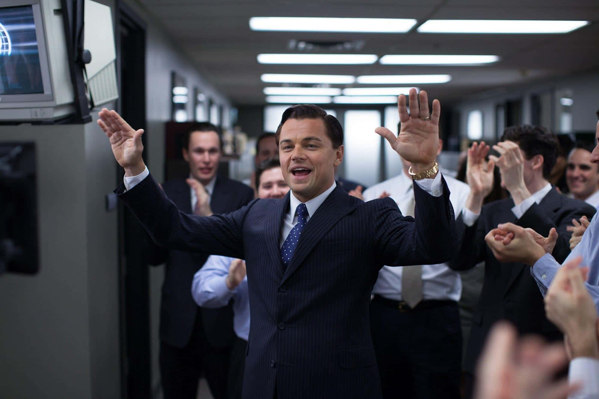 Wolf Of Wall Street 桌布 壁紙 — 李奧納多·狄卡皮歐身穿細條紋西裝，在公司辦公室的走廊高舉雙臂慶祝勝利，同事們在周圍歡呼。