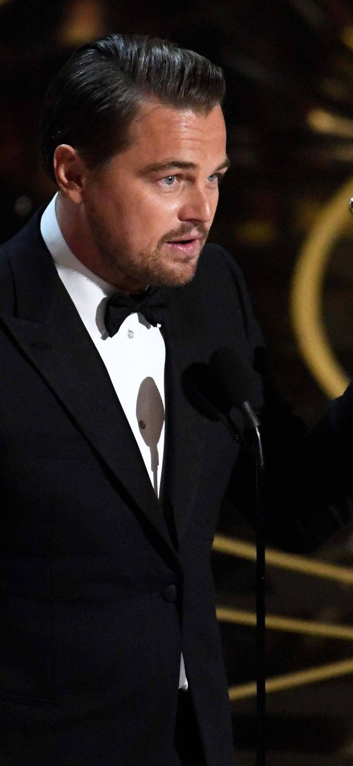 Dicaprio 桌布 壁紙 — 李奧納多·狄卡皮歐身穿黑色禮服，在溫暖的金色舞台燈光下於正式頒獎典禮上發言。