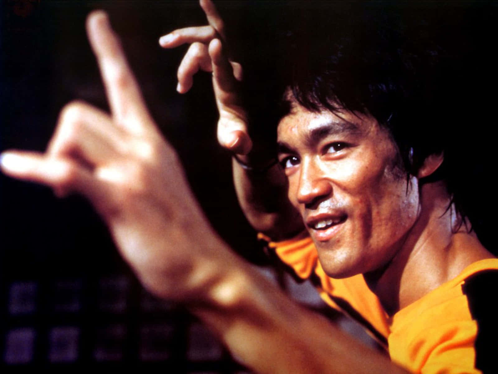 李小龍 桌布 壁紙 — 李小龍以動感武術姿勢，身穿橙色襯衫，背景深色。