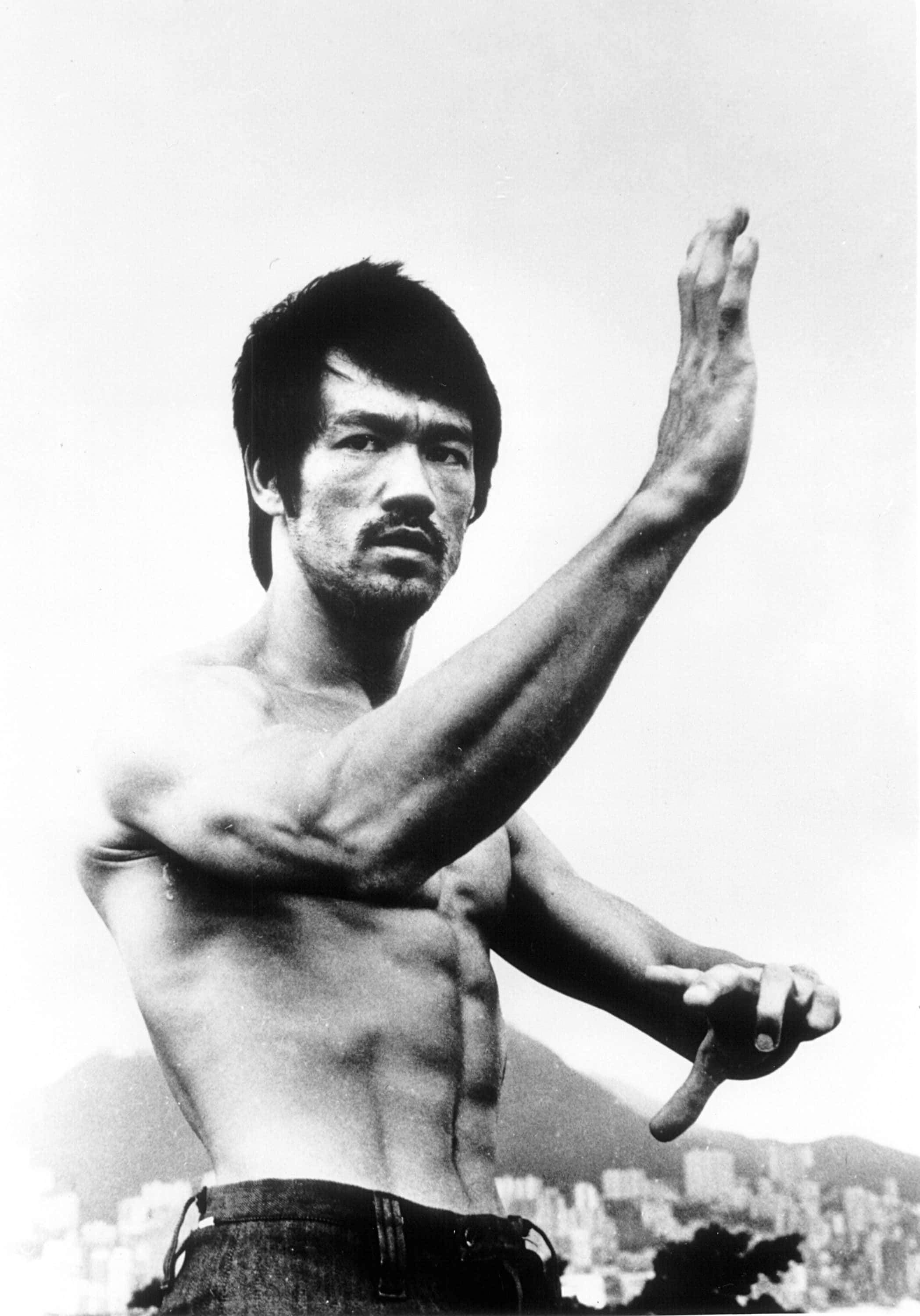李小龍 桌布 壁紙 — 李小龍以強勁武術姿勢赤裸上身，背景是模糊的城市天際線。