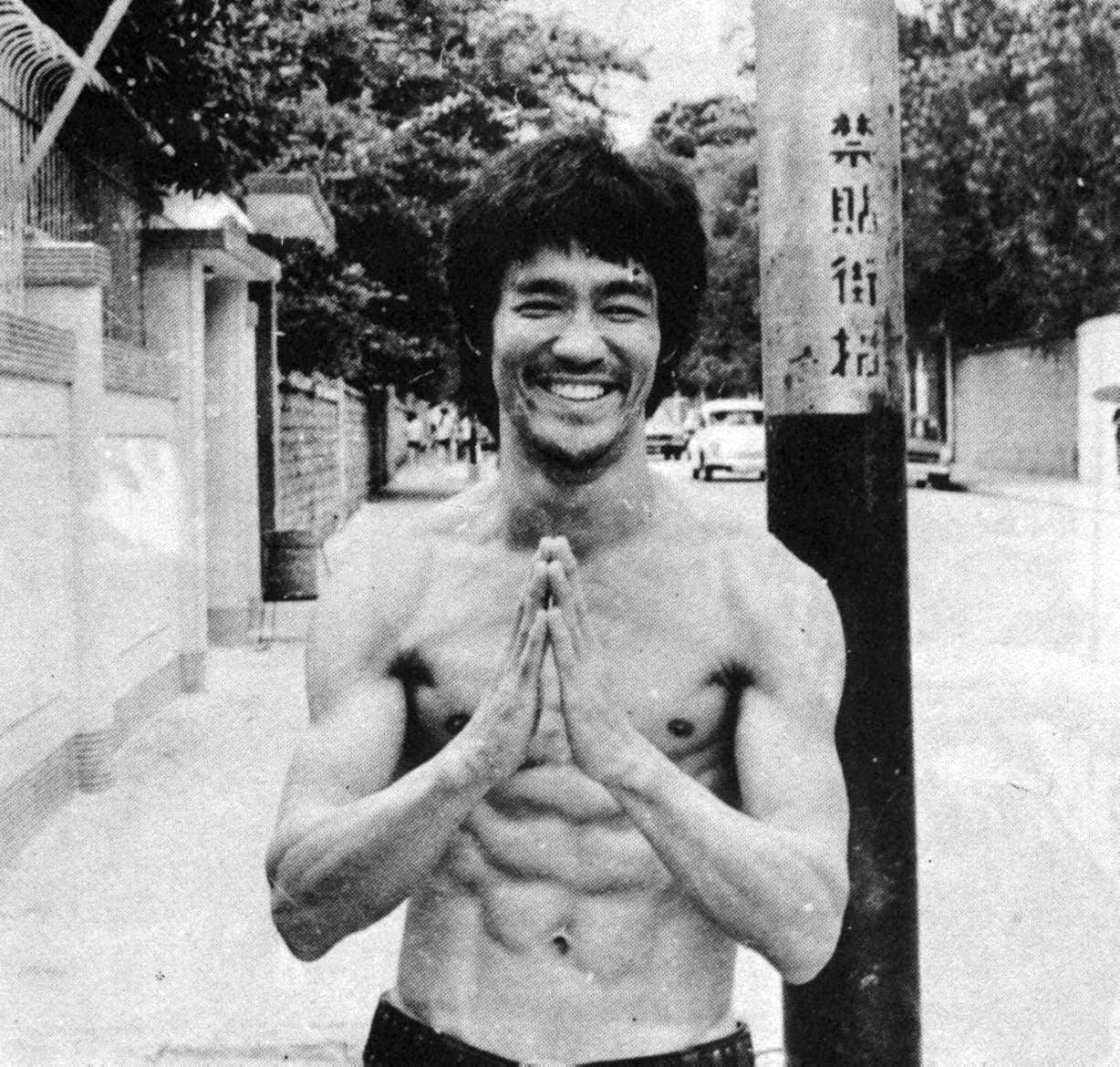 李小龍 桌布 壁紙 — 李小龍在陽光普照的城市小巷中，赤膊雙手合十，在街燈柱前溫暖地微笑。