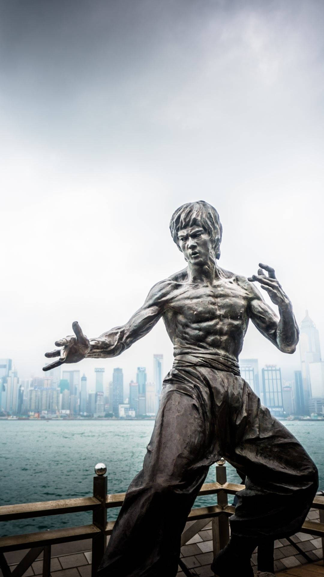 李小龍 桌布 壁紙 — 李小龍的青銅雕像以動態武術姿勢矗立在海濱碼頭上，背後是朦朧的城市天際線。