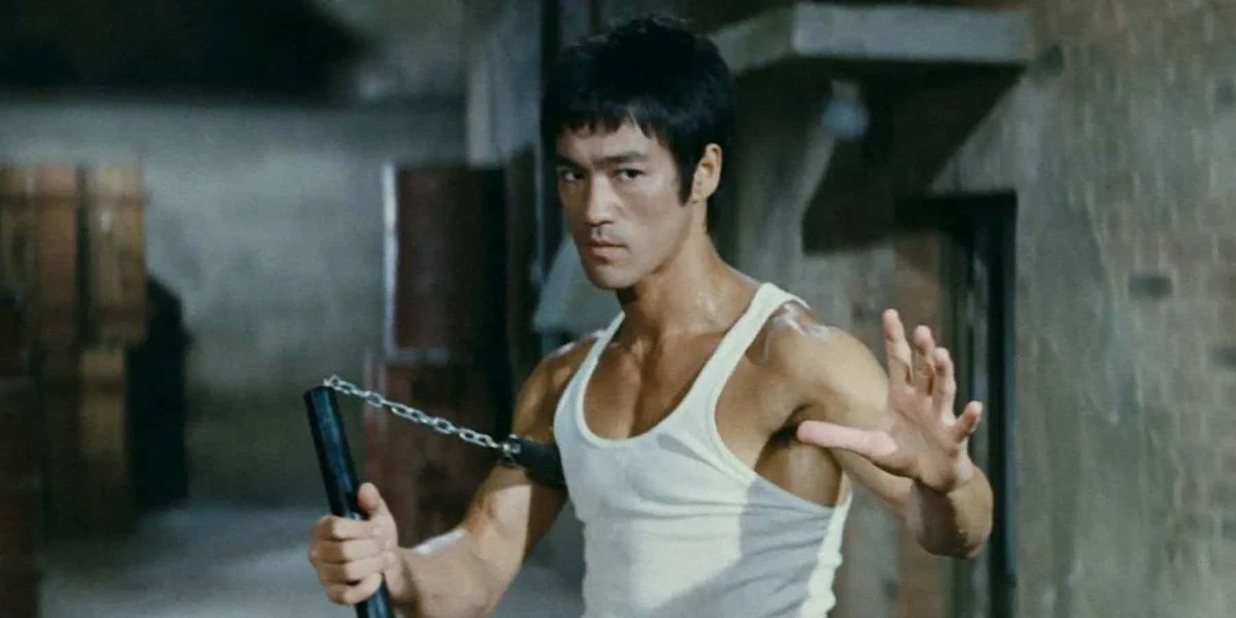 李小龍 桌布 壁紙 — 李小龍站在一個骯髒的倉庫裡，手持雙節棍，神情專注，肌肉線條分明。