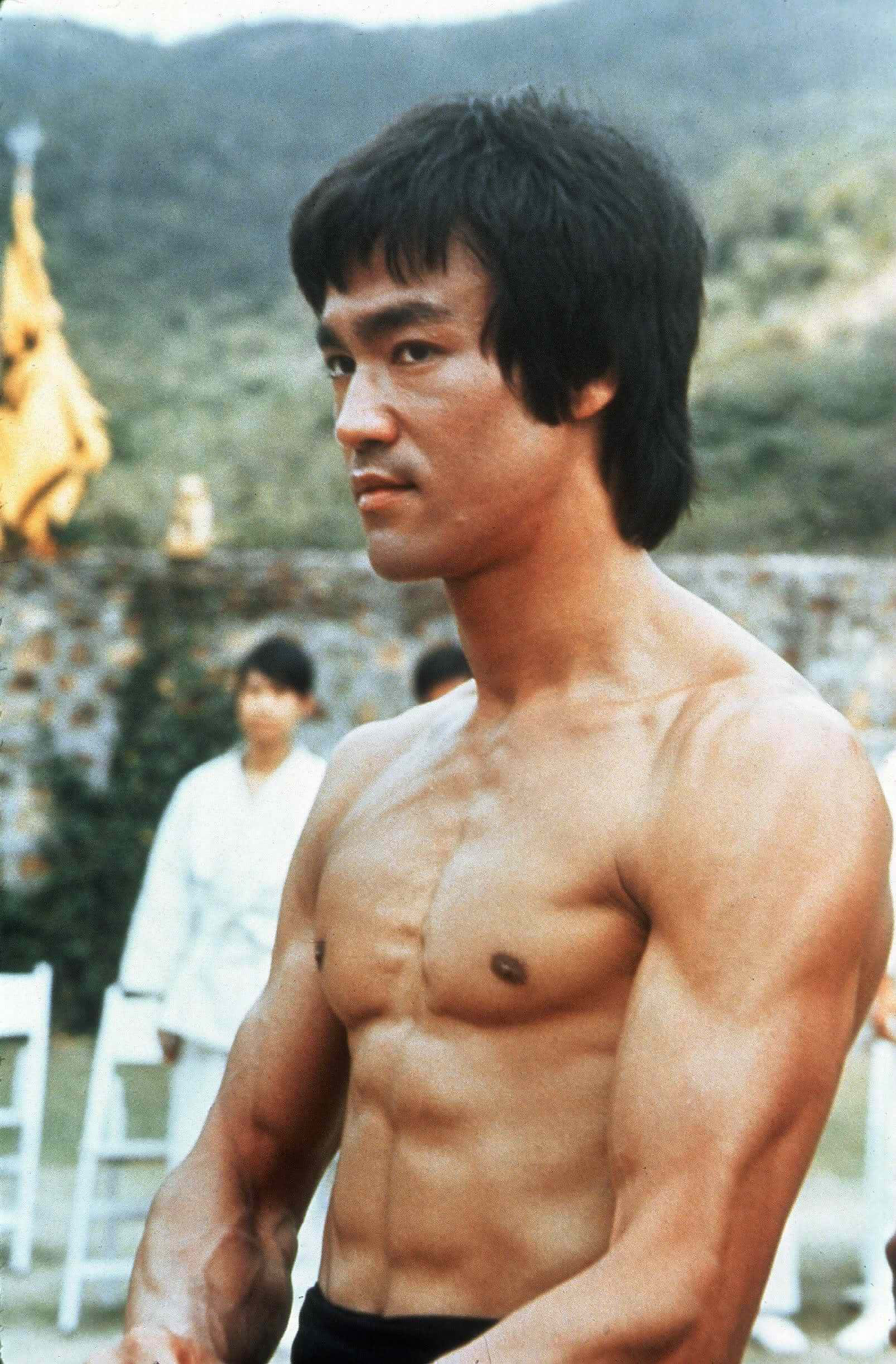 李小龍 桌布 壁紙 — 李小龍赤裸上身，展現出清晰的肌肉線條，背景是山脈，有穿著白色服裝的模糊人物。