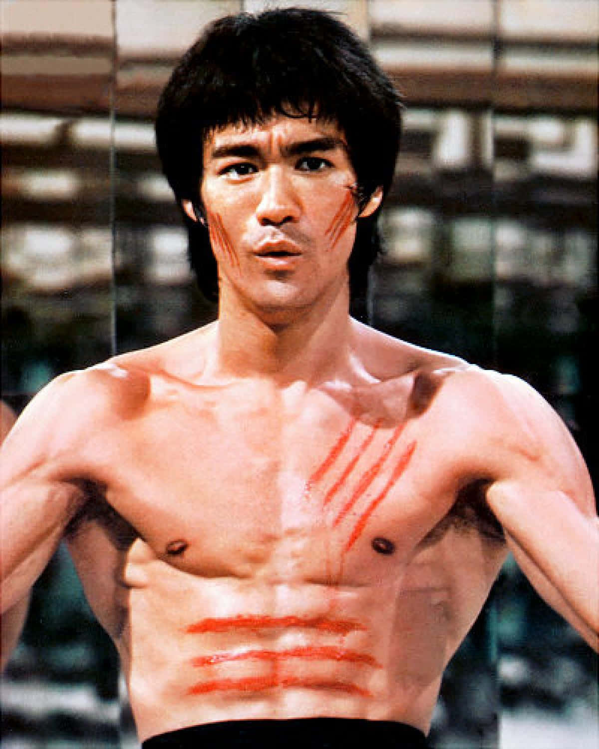李小龍 桌布 壁紙 — 李小龍赤裸上身，胸口和臉頰上有紅色劃痕，背景是模糊的城市景觀，眼神專注。
