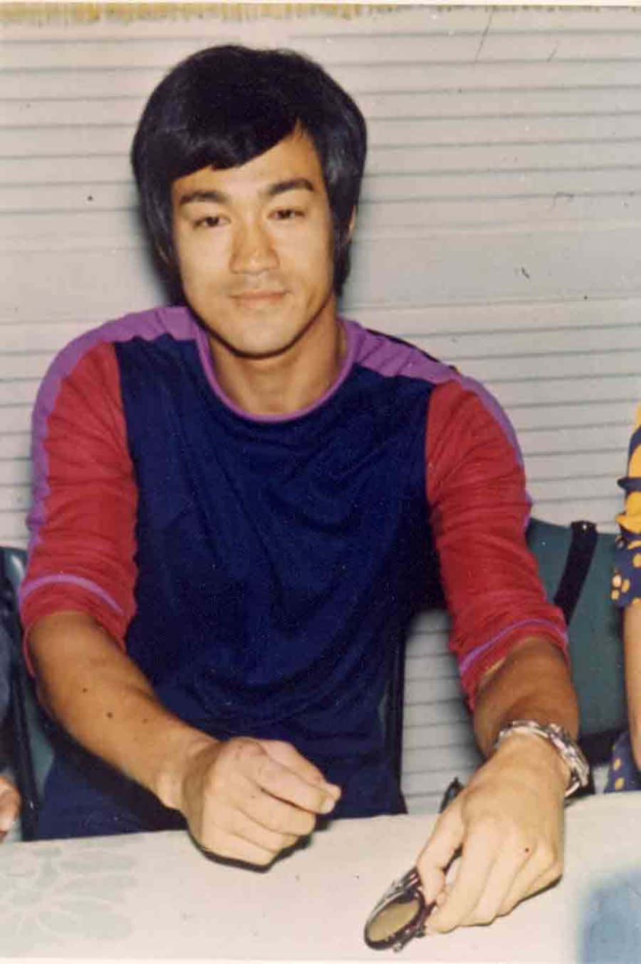 李小龍 桌布 壁紙 — 李小龍身穿紅藍衫，靜靜坐在桌前，手持眼鏡的復古照片。