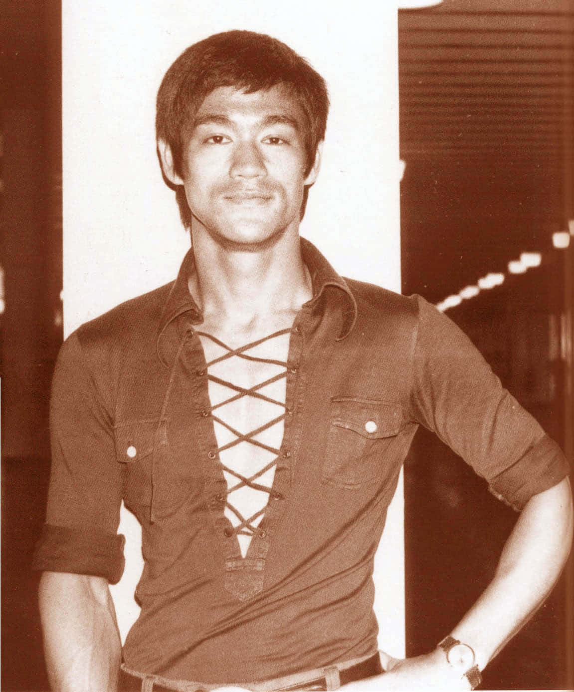 李小龍 桌布 壁紙 — 李小龍身穿繫帶襯衫，在昏暗背景中帶著溫暖的懷舊色調自信微笑。