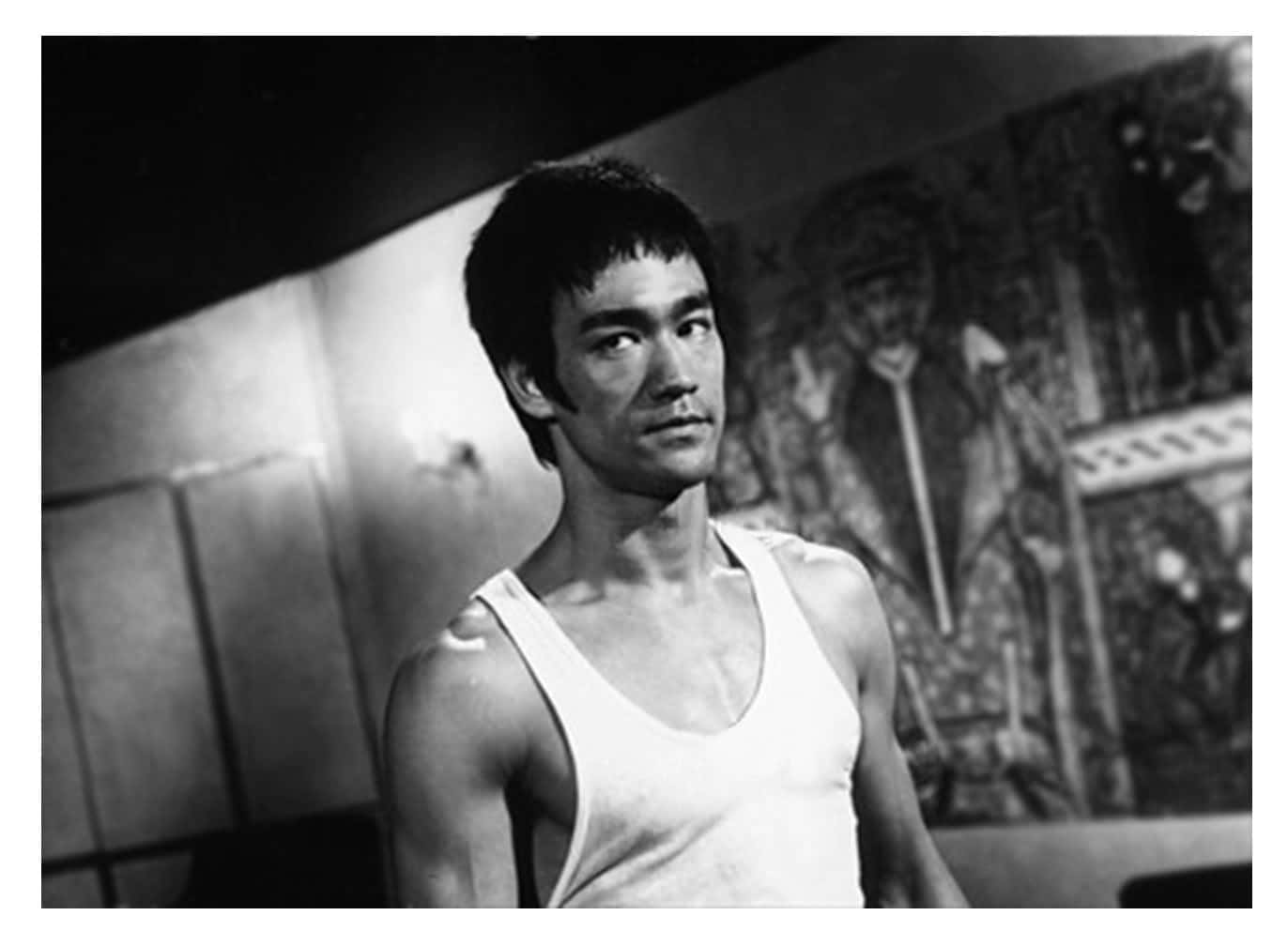 李小龍 桌布 壁紙 — 李小龍身處燈光昏暗的房間，身穿白色背心，背後有壁畫，經典黑白照片。