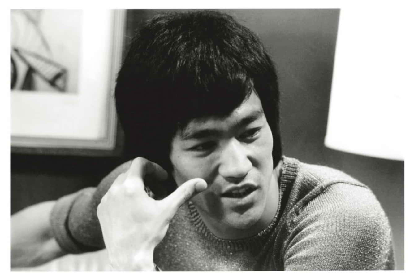 李小龍 桌布 壁紙 — 李小龍黑白照片，沉思表情，手靠近臉部，室內環境有燈和裱框藝術品。