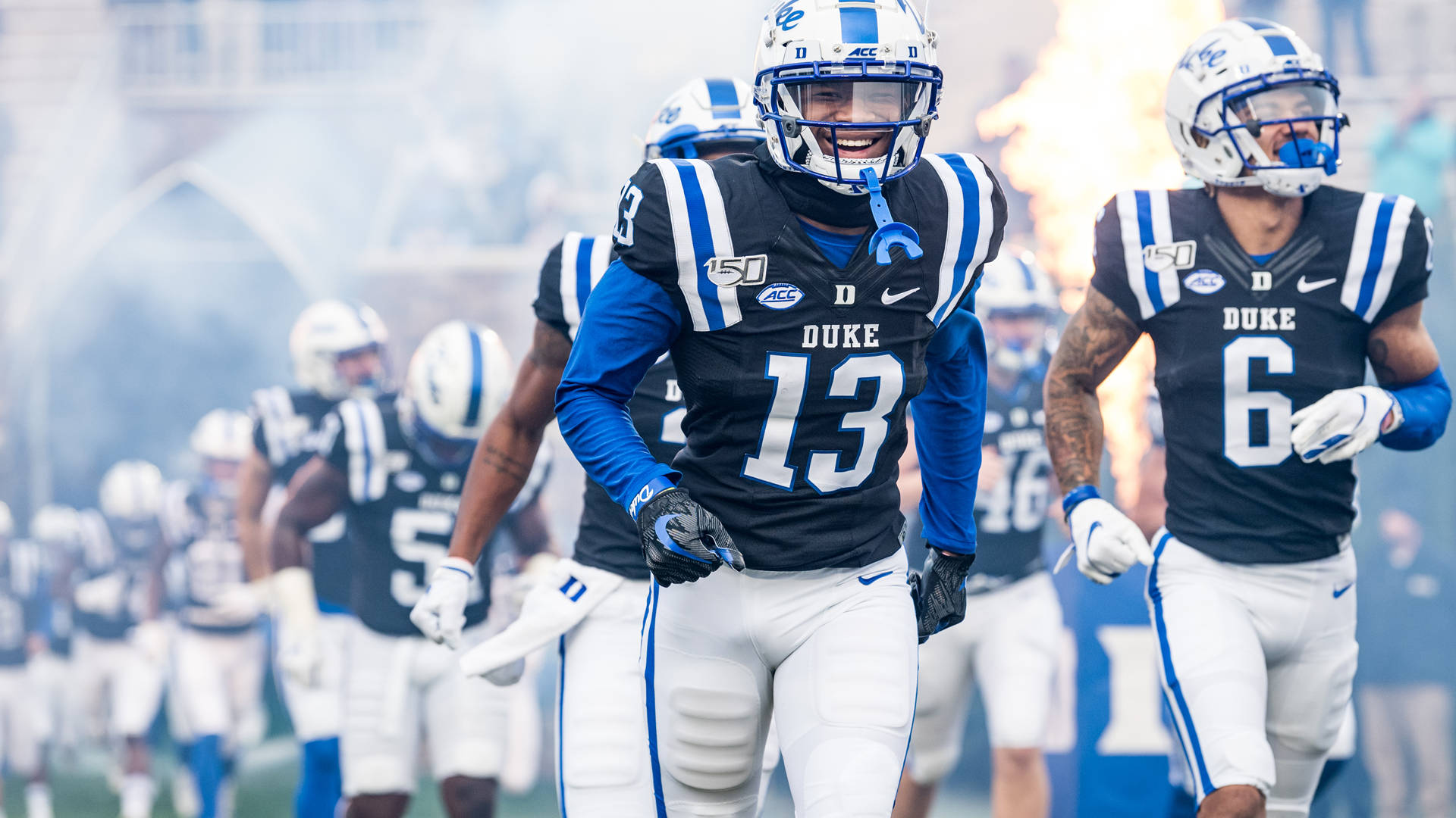 Duke Blue Devils 桌布 壁紙 — 杜克大學美式足球員身穿藍黑制服，穿過煙霧和火焰效果跑入球場。