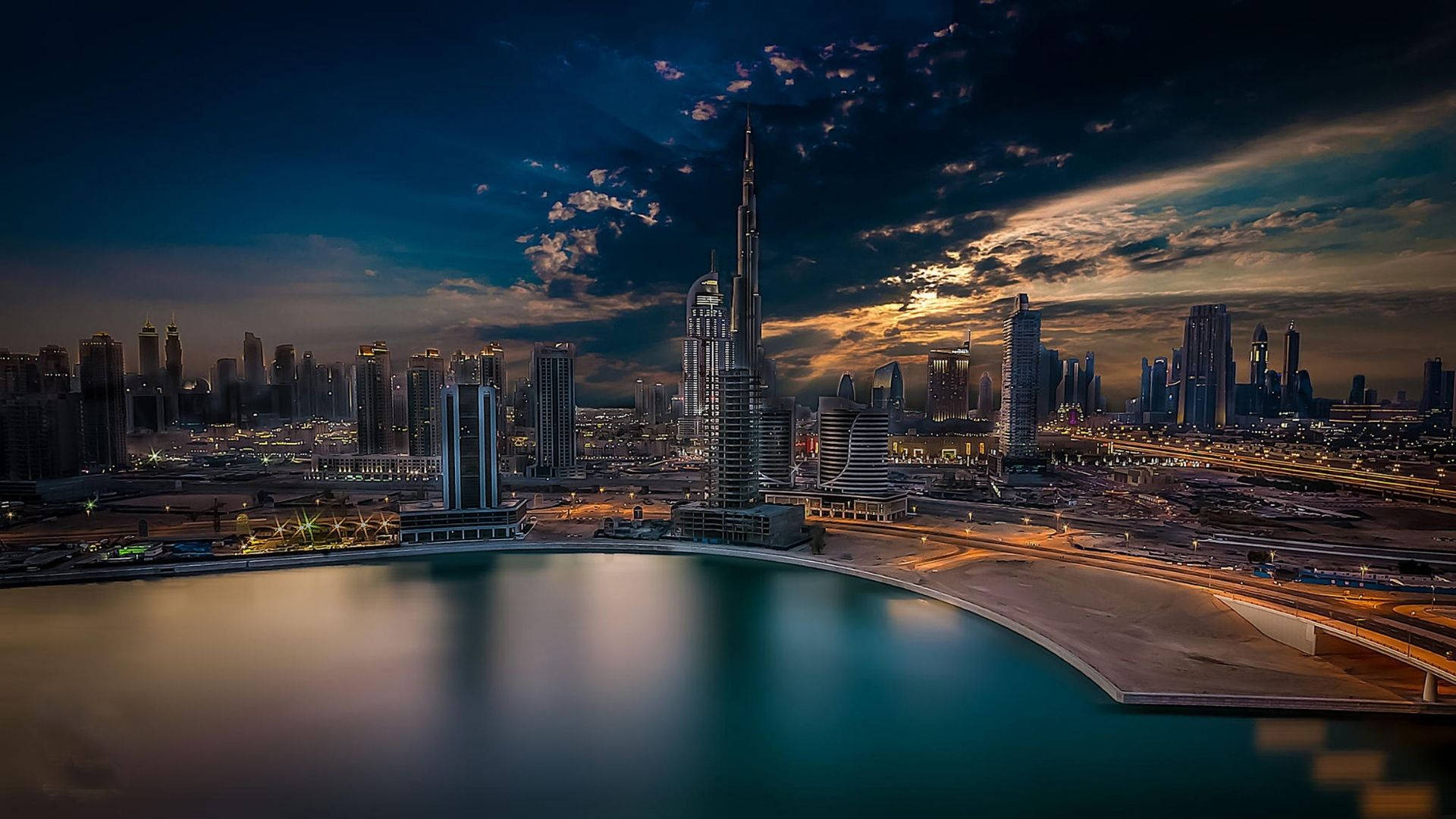 Dubai 桌布 壁紙 — 杜拜令人驚嘆的黃昏城市景觀，哈里發塔聳立在閃爍的摩天大樓之上，平靜的水面倒映著戲劇性的天空。