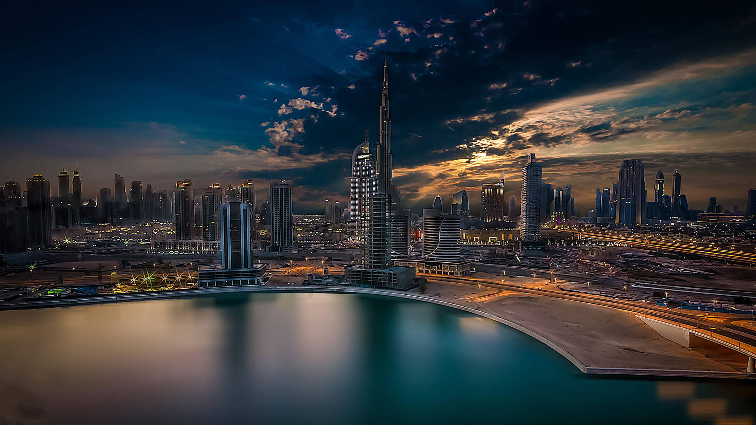 Arab 桌布 壁紙 — 杜拜令人驚嘆的黃昏城市景觀，哈里發塔聳立在閃爍的摩天大樓之上，寧靜的海濱倒映著戲劇性的天空。