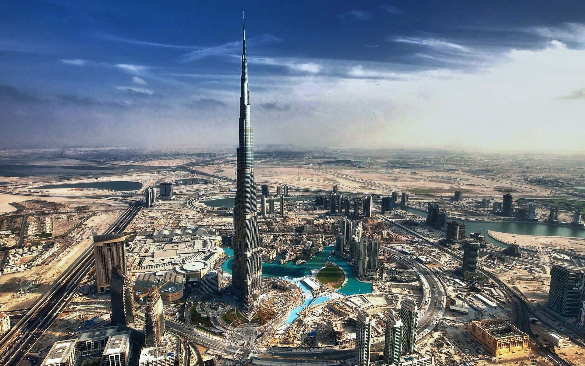 Uae 桌布 壁紙 — 杜拜天際線的航拍圖，哈里發塔聳立於現代摩天大樓和沙漠景觀之上，背景是戲劇性的藍天。