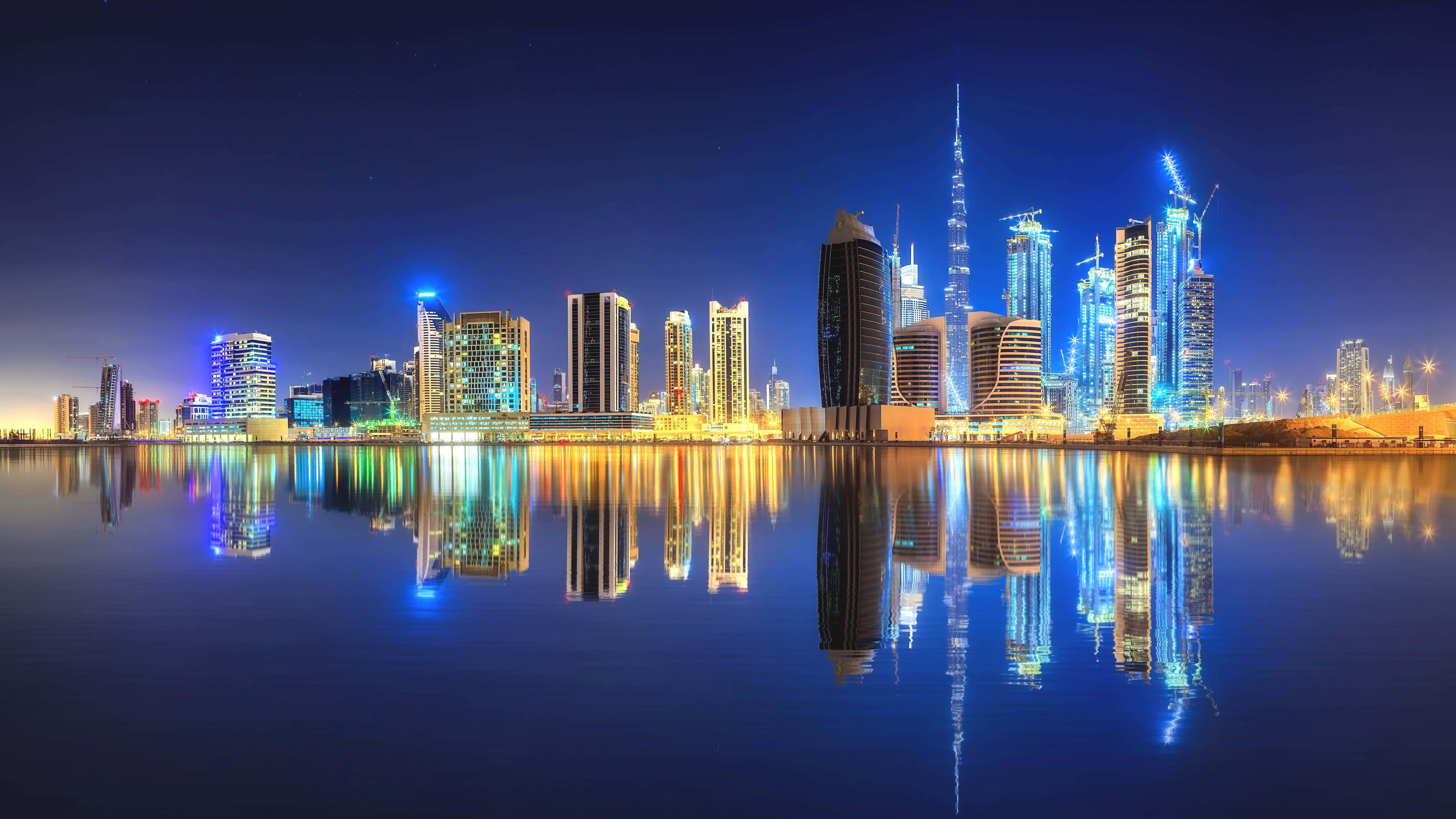Uae 桌布 壁紙 — 杜拜天際線的迷人夜景，摩天大樓閃爍著藍色和金色光芒，完美倒映在平靜的水面上。
