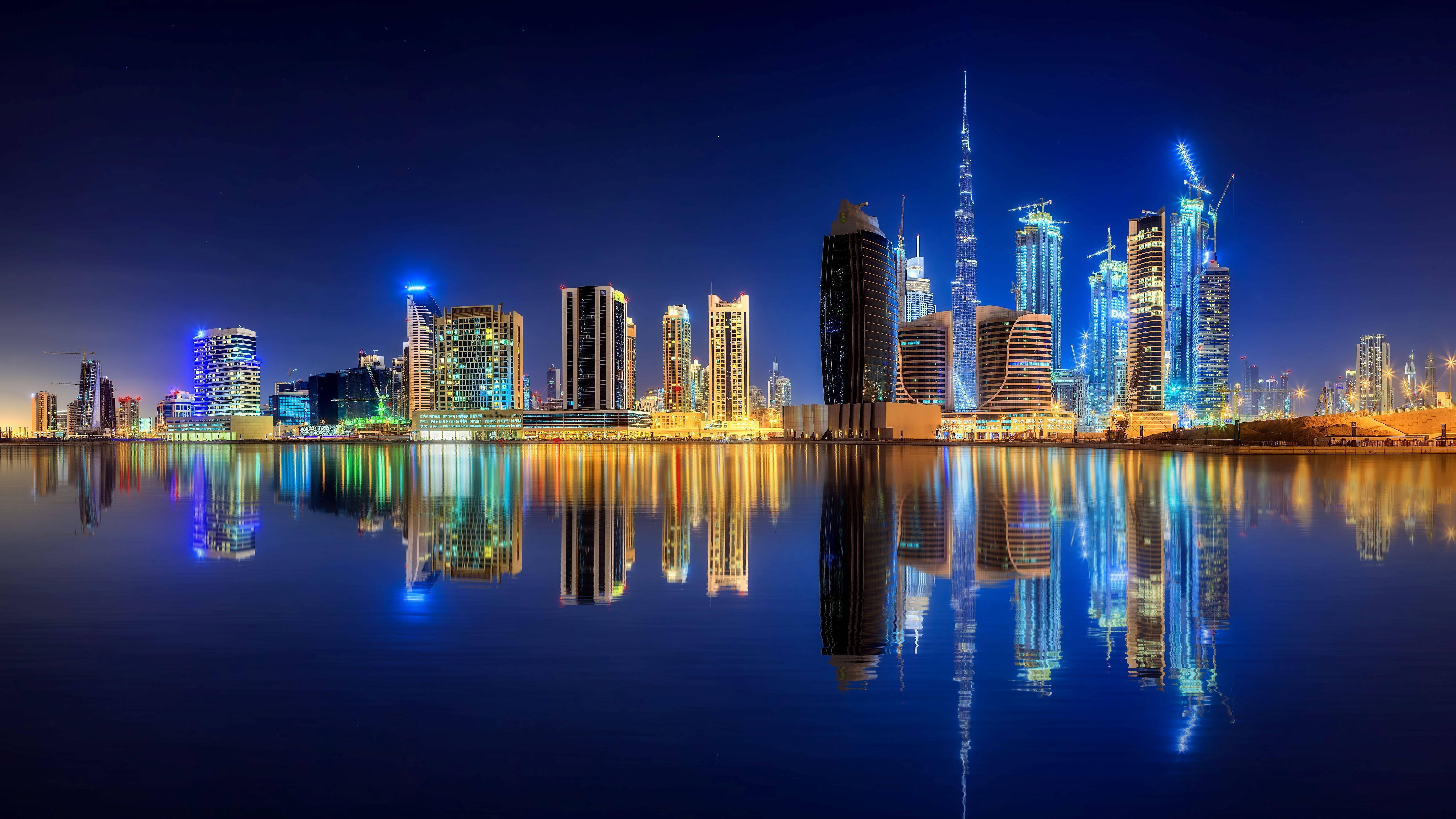 Arab 桌布 壁紙 — 杜拜天際線的迷人夜景，摩天大樓閃爍著藍色和金色光芒，完美倒映在平靜的水面上。