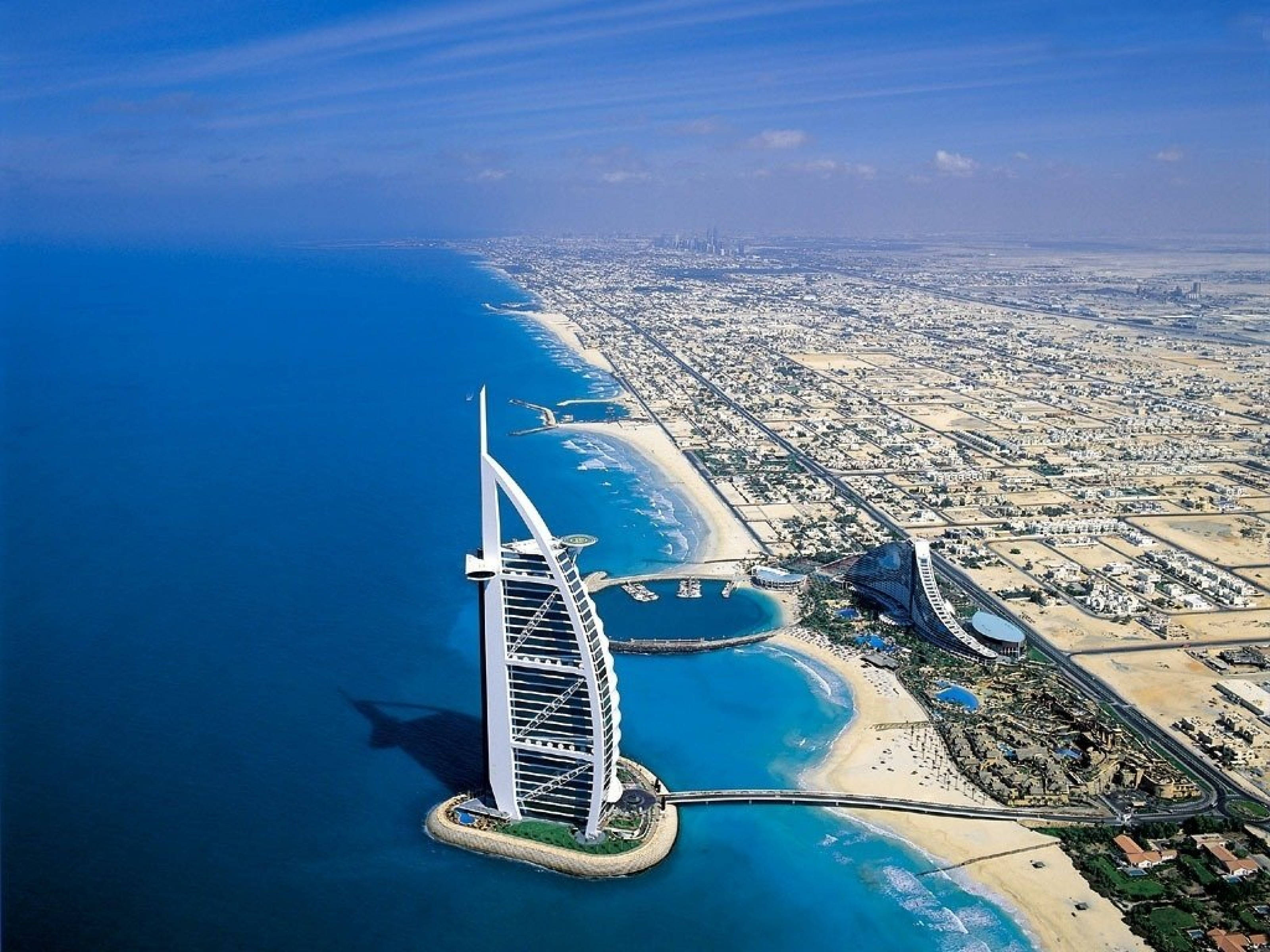 風景動畫 桌布 壁紙 — 杜拜標誌性帆船飯店（Burj Al Arab）的鳥瞰圖，飯店獨自矗立於人工島上，四周是碧綠的海水，內陸延伸著沙漠城市景觀。