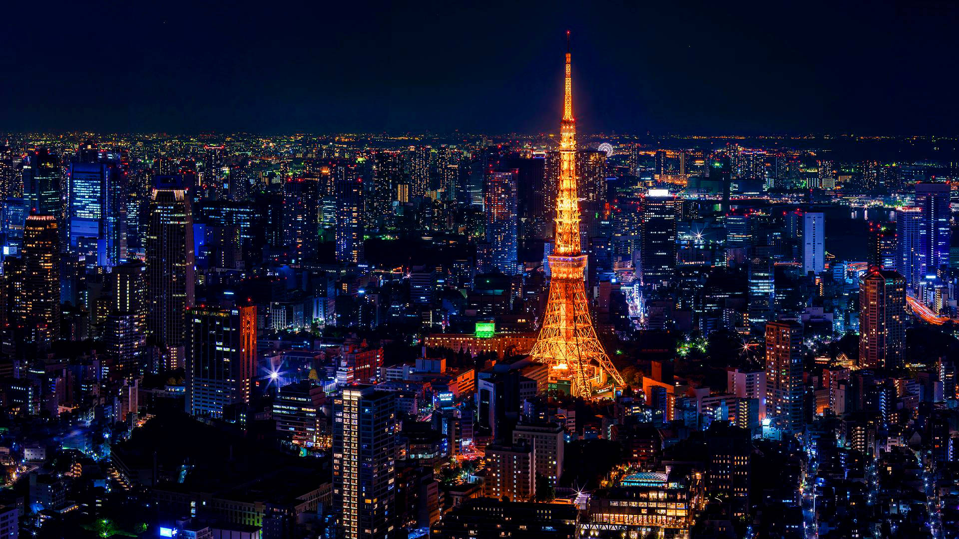 日本 桌布 壁紙 — 東京塔在黑暗的天空中溫暖地發光，照亮了夜晚藍色和橙色霓虹燈蔓延的城市景觀。