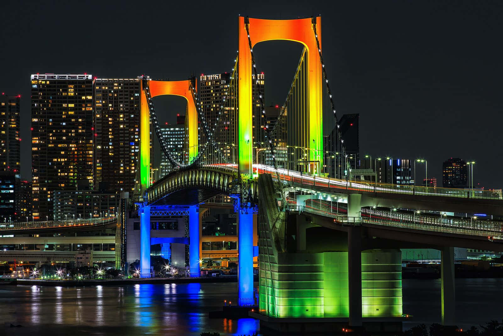 Rainbow Bridge 桌布 壁紙 — 東京彩虹橋在漆黑的夜空下閃爍著鮮豔的彩虹燈光，倒映在水面上，背後是城市的高樓大廈。