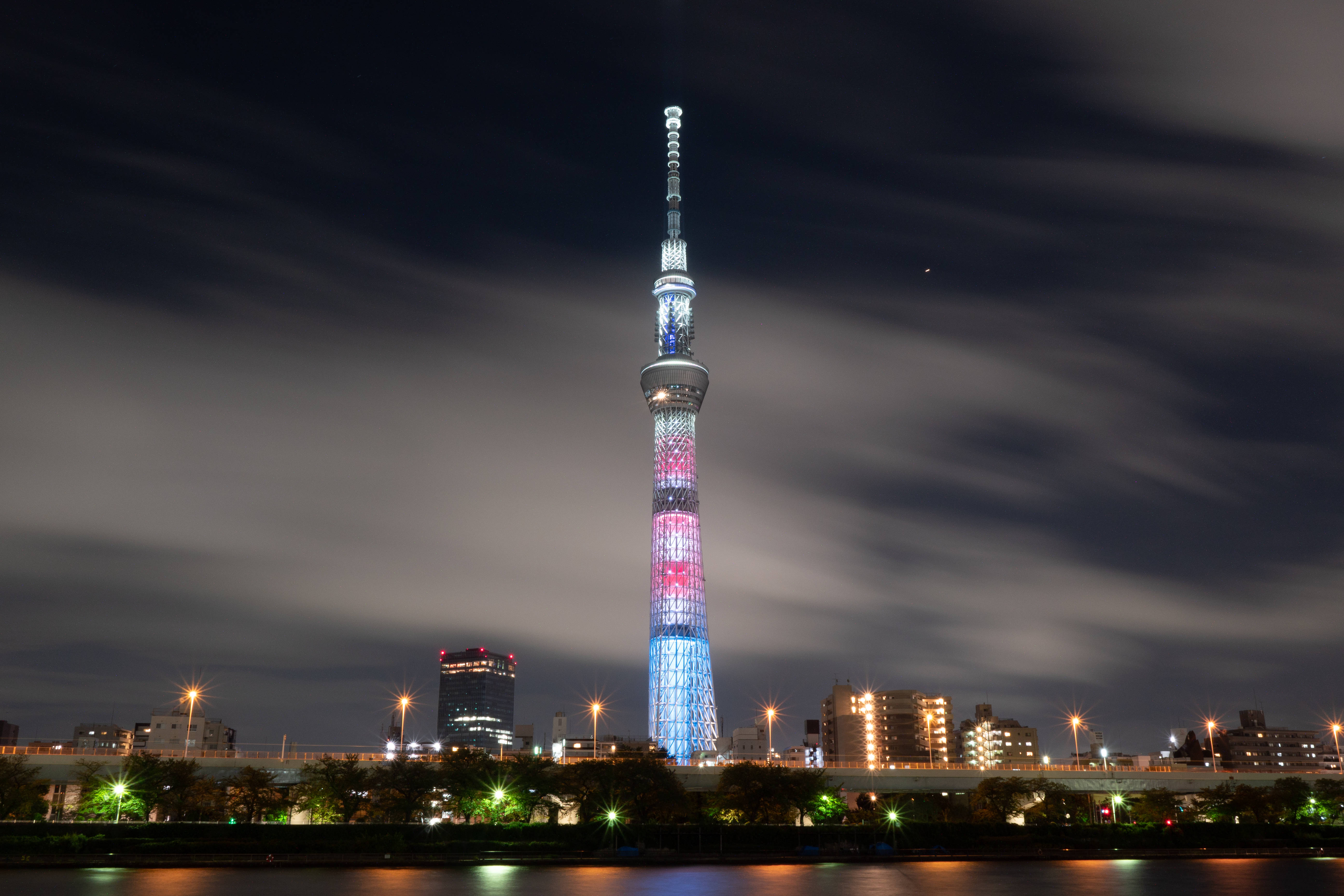 Architecture 桌布 壁紙 — 東京晴空塔在漆黑的夜空中被鮮豔的燈光照亮，有條紋狀的雲朵和城市燈光倒映在下面的水面上。
