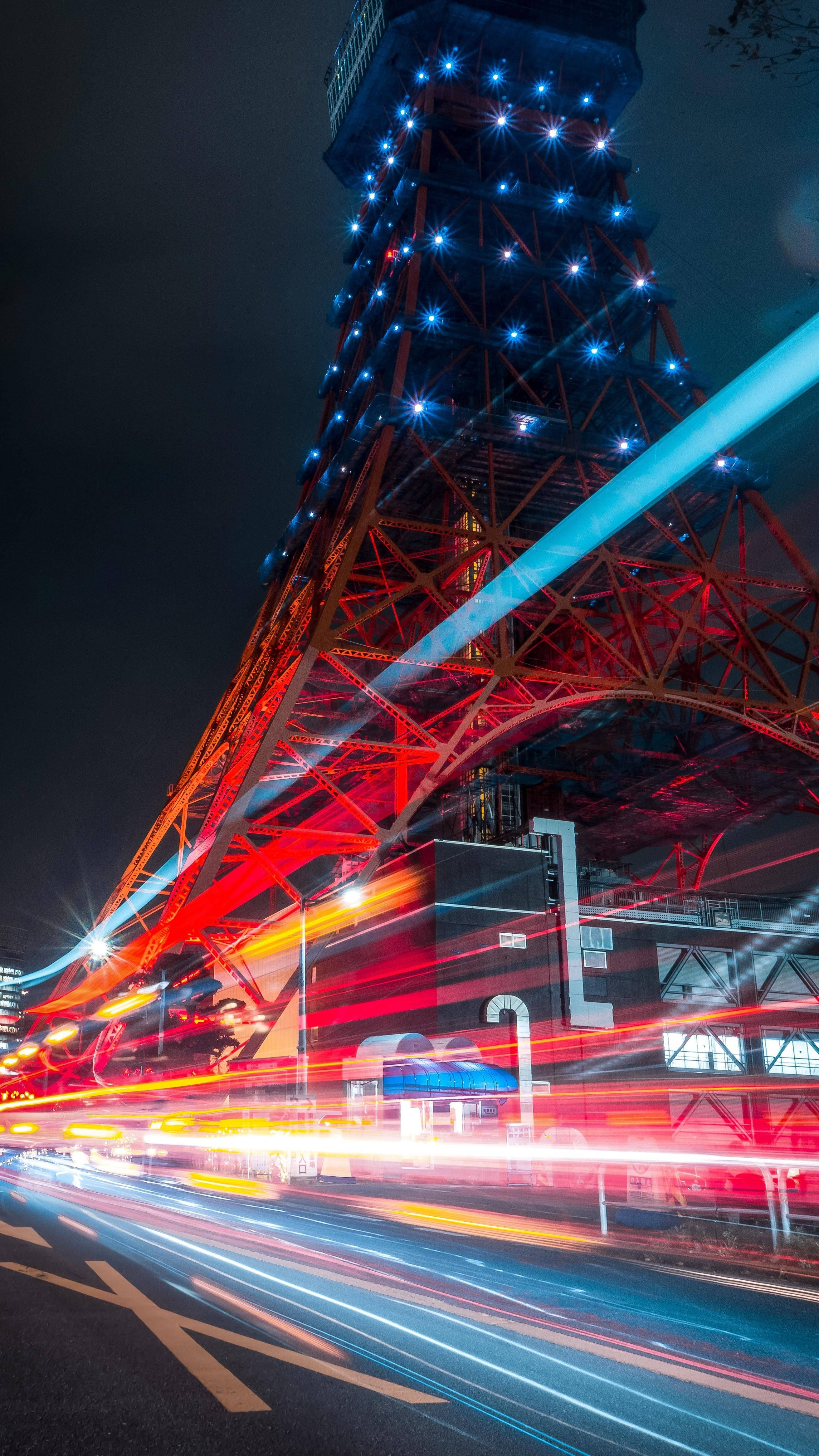 日本 桌布 壁紙 — 東京鐵塔在夜晚閃耀紅藍光芒，下方車流形成光軌的充滿活力的夜景照片。