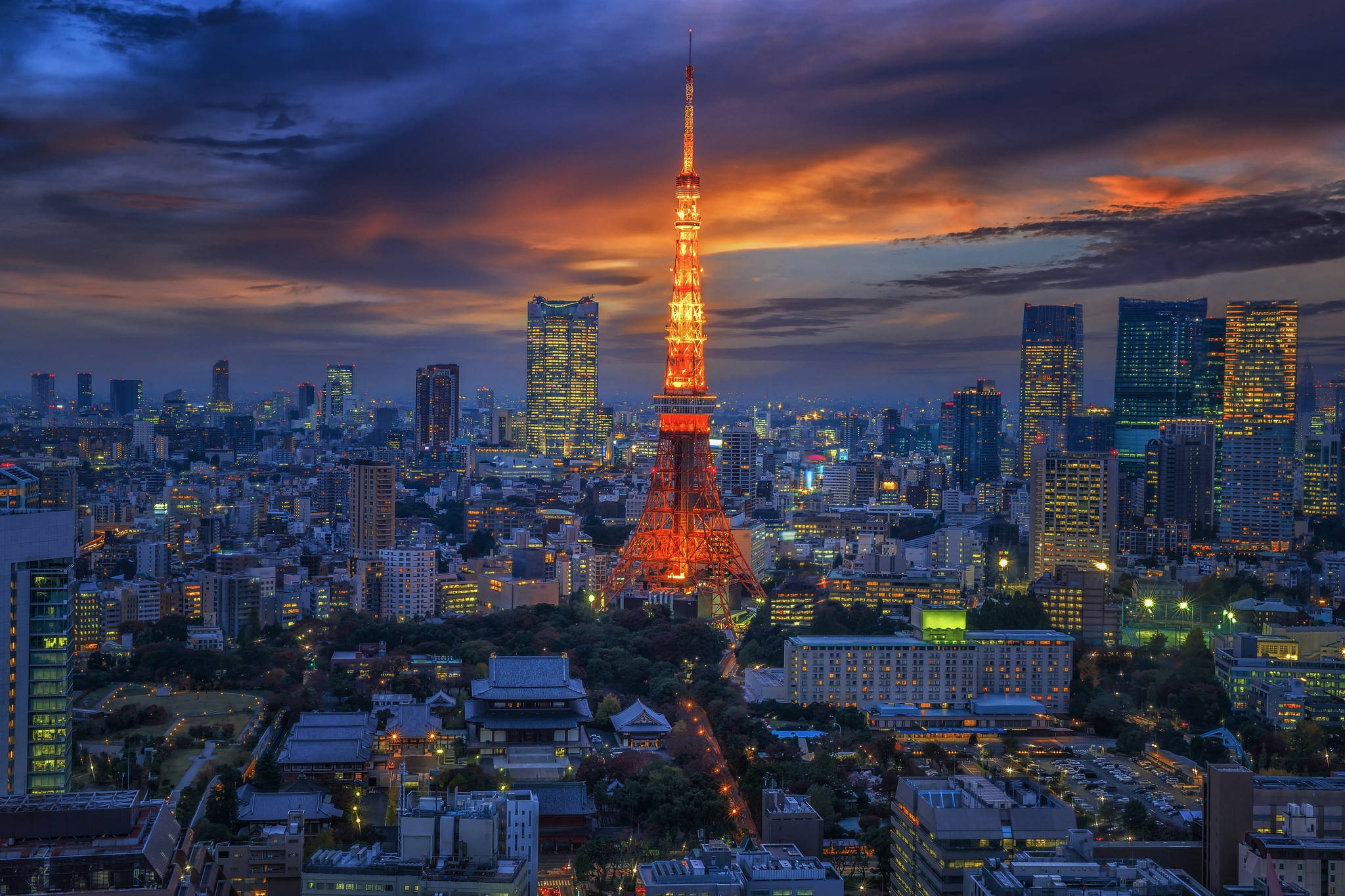 日本 桌布 壁紙 — 東京鐵塔在戲劇性的暮色天空中溫暖地發光，城市燈光照亮了下面的城市擴展區。