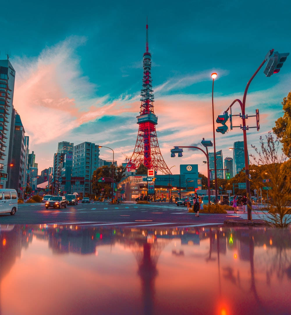 日本 桌布 壁紙 — 東京鐵塔在暮色天空下發出紅光，倒映在濕漉漉的城市街道上，有汽車和行人。
