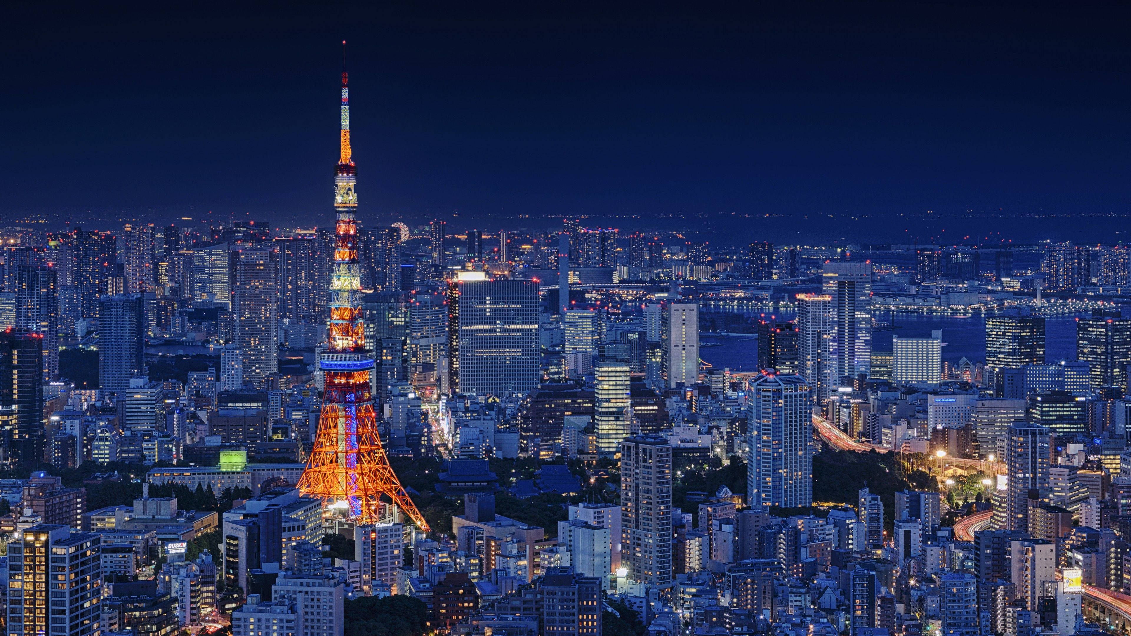 日本 桌布 壁紙 — 東京鐵塔在深藍色的夜空中發出橙色光芒，下方是廣闊的城市景觀，佈滿了燈火通明的摩天大樓和街道。