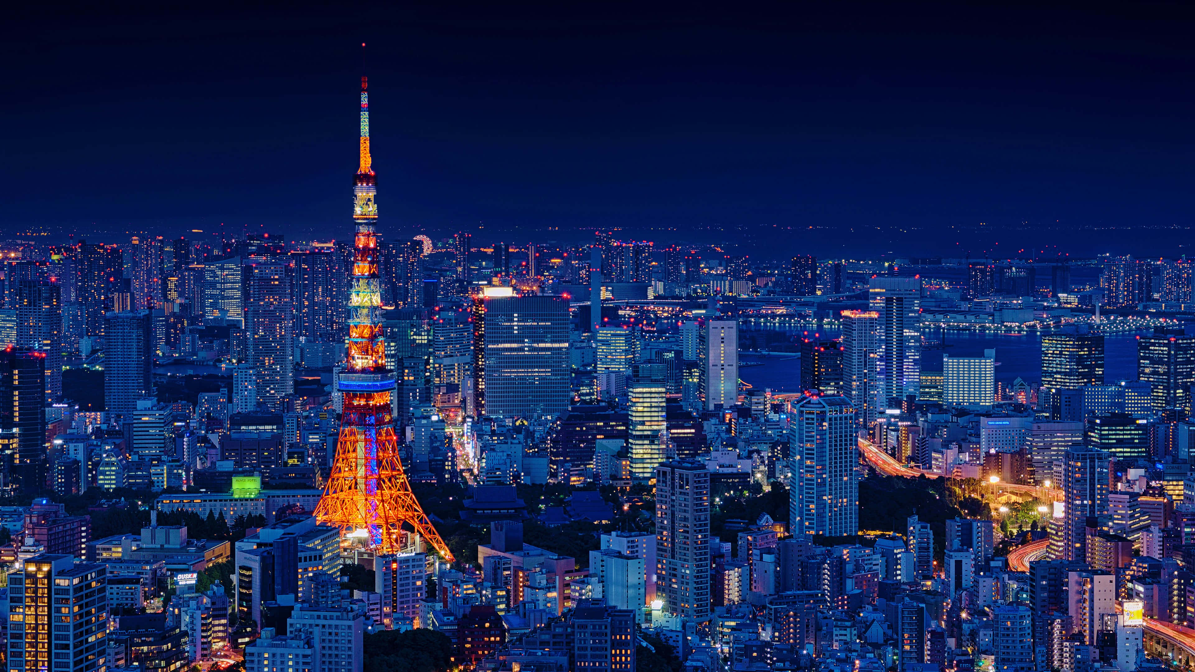東京鐵塔 桌布 壁紙 — 東京鐵塔在深藍色的夜空中發出鮮豔的橙色光芒，下方是廣闊的城市景觀，閃爍的燈光和高聳的摩天大樓。