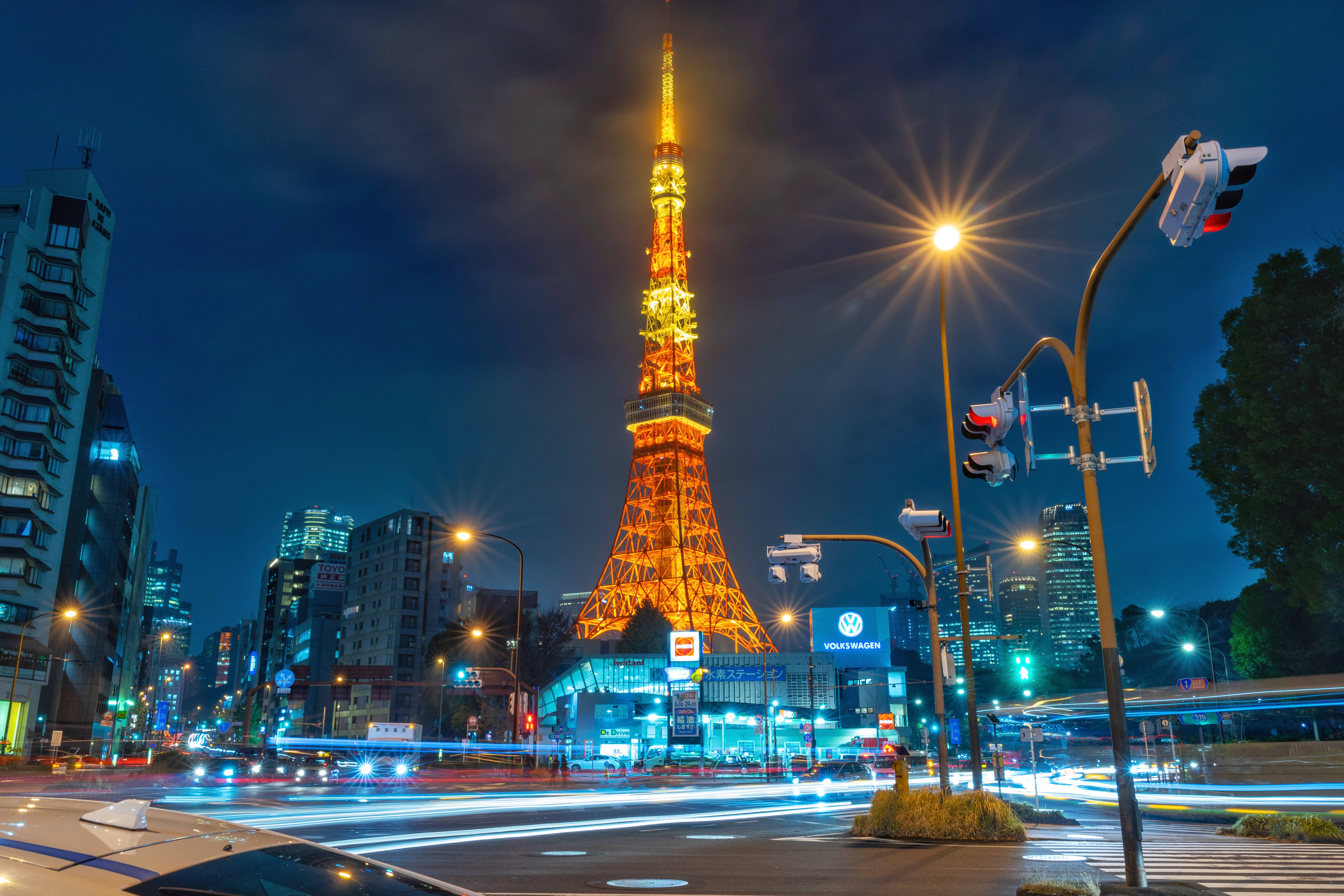 東京鐵塔 桌布 壁紙 — 東京鐵塔在深藍色的夜空中閃耀著金色光芒，下方繁忙的城市街道上，車燈的軌跡模糊地掠過。