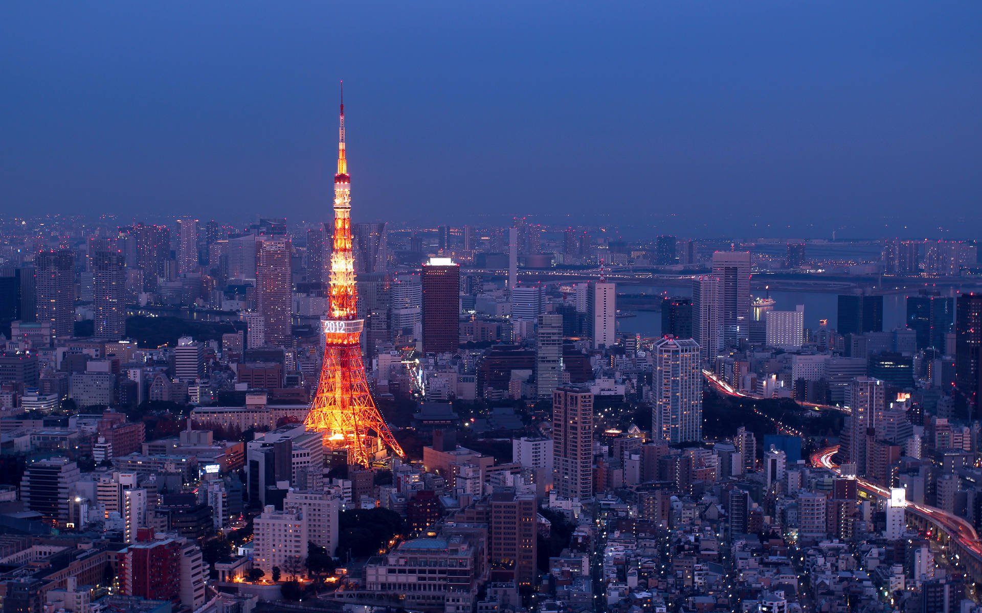 日本 桌布 壁紙 — 東京鐵塔在深藍色的暮色天空中溫暖地發光，下方是廣闊的城市景觀，街道和建築物閃爍著光芒。