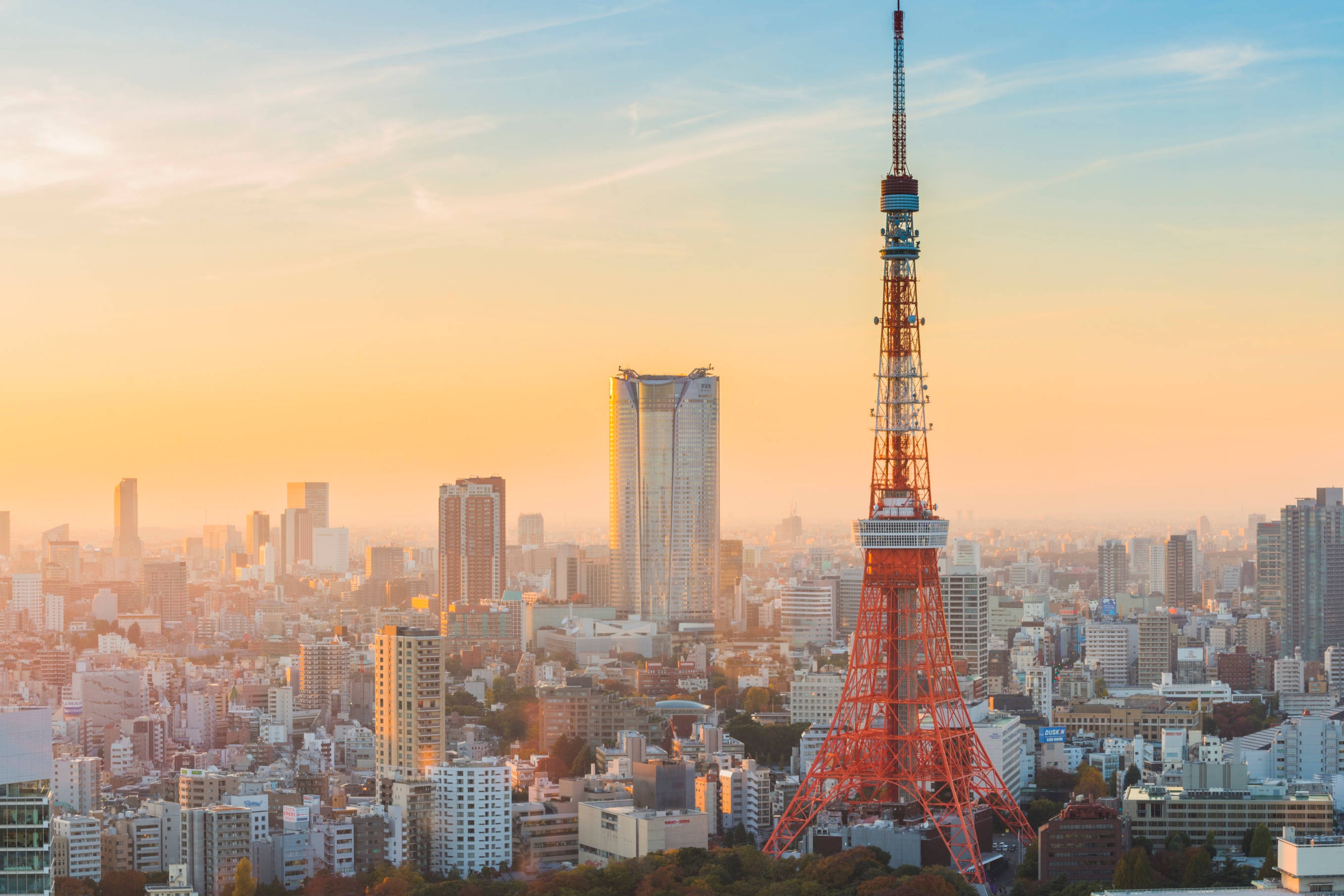 日本 桌布 壁紙 — 東京鐵塔在金色夕陽天空下，聳立於現代摩天大樓和低層建築組成的廣闊城市天際線之上。
