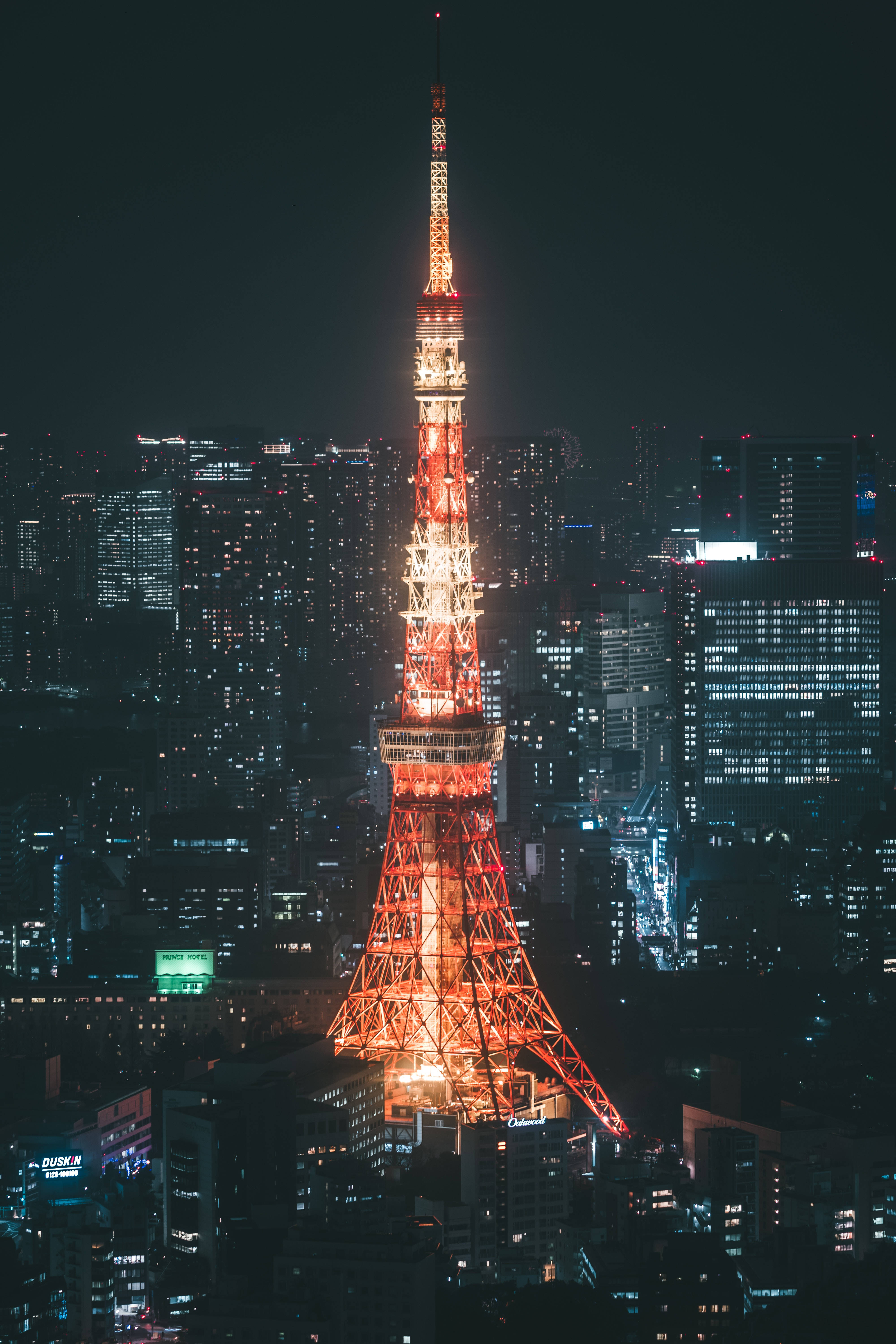 日本 桌布 壁紙 — 東京鐵塔在黑暗的天空中溫暖地發光，周圍是夜晚燈火輝煌的摩天大樓閃爍的城市景觀。