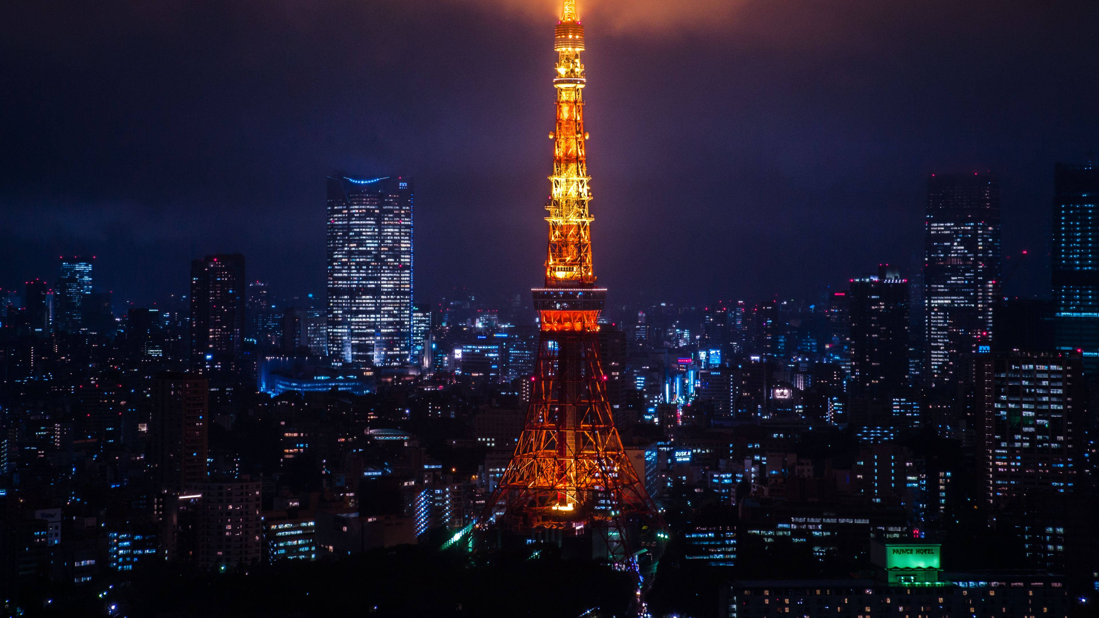日本 桌布 壁紙 — 東京鐵塔在黑暗的天空中溫暖地發光，城市燈光在下方閃爍，構成一個充滿活力的夜間大都會。