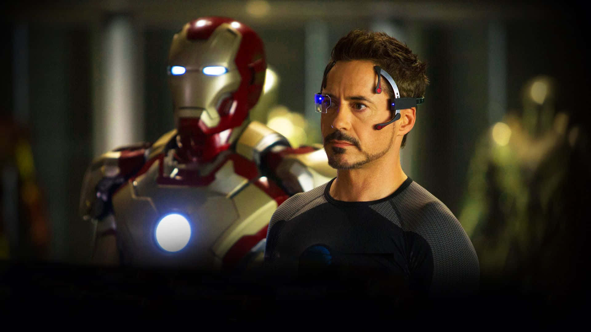 Iron Man Movies 桌布 壁紙 — 東尼·史塔克在他的實驗室中，身後有發光的鋼鐵人戰衣，處於昏暗的未來感燈光下。