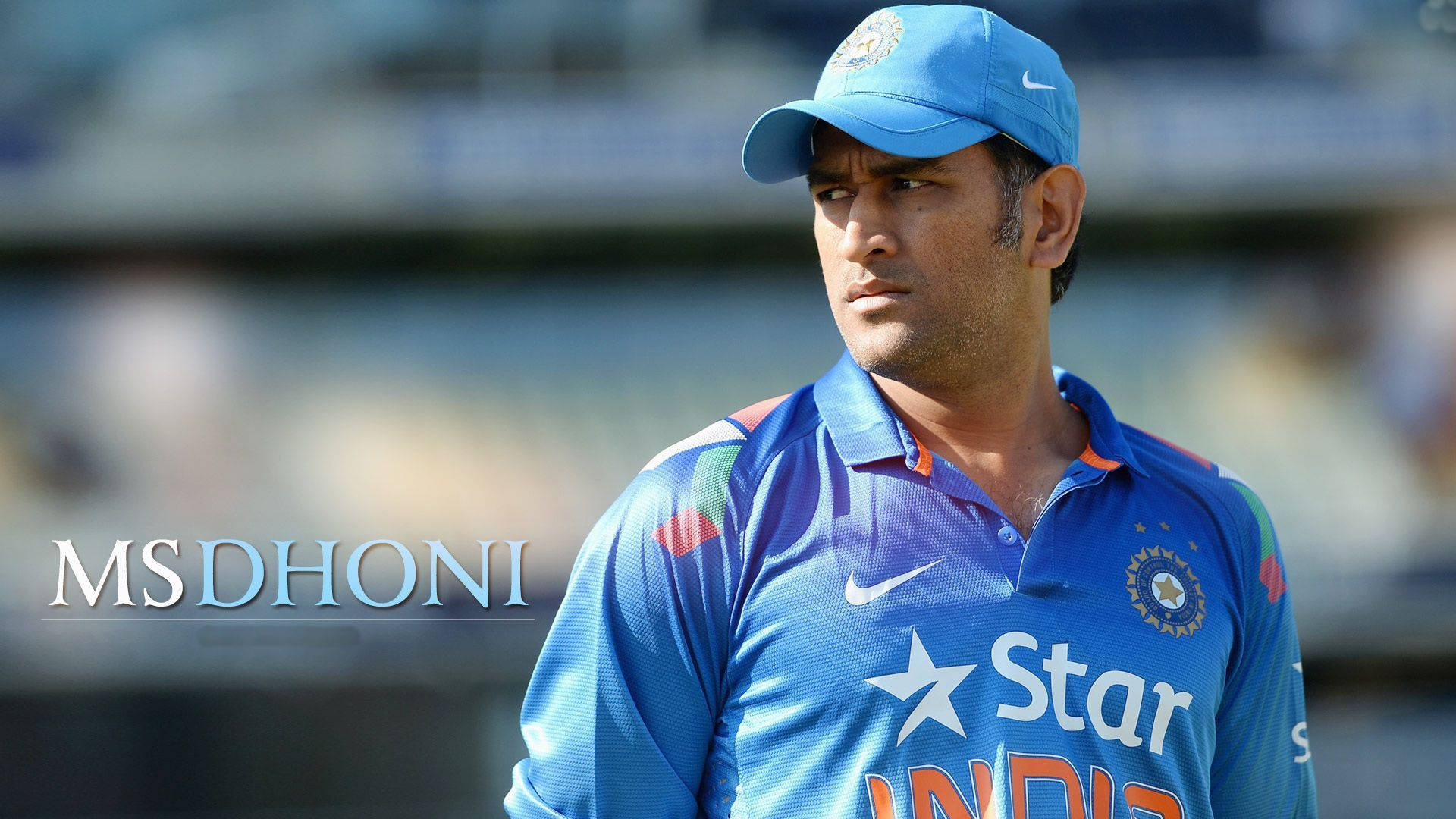Ms Dhoni 桌布 壁紙 — 板球傳奇人物MS Dhoni身穿藍色印度球衣，在日間比賽中專注凝視，背景球場模糊。