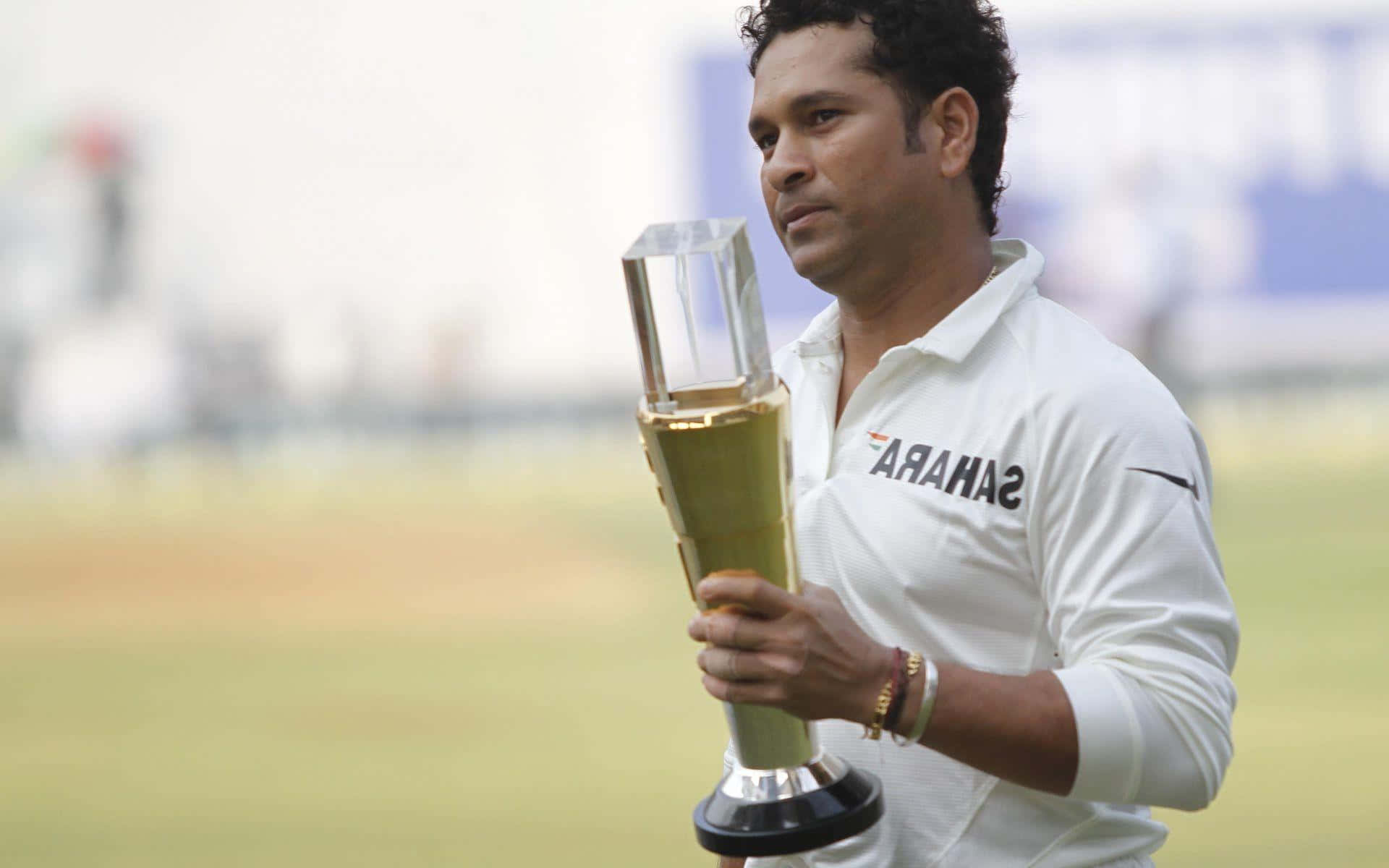 Sachin Tendulkar 桌布 壁紙 — 板球傳奇人物沙欽·坦杜卡在陽光明媚的綠色球場上，神情專注地手持閃閃發光的金色獎盃。