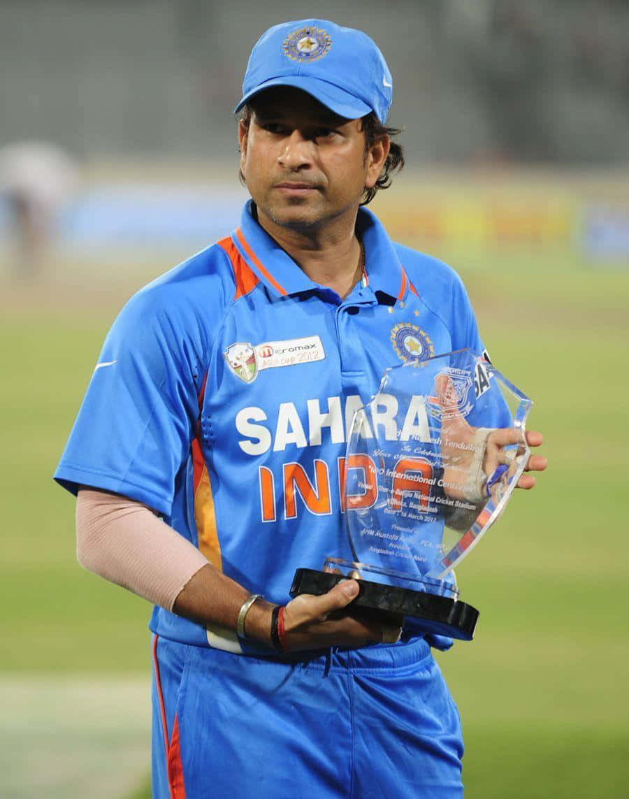 Sachin Tendulkar 桌布 壁紙 — 板球傳奇沙欽·坦杜卡穿上藍色印度球衣，在球場燈光下的綠色草地上手持玻璃獎盃。