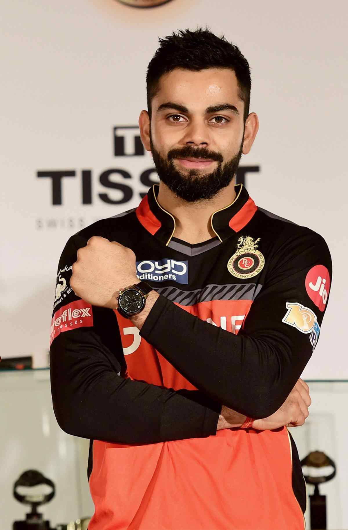 virat kohli ipl 桌布 壁紙 — 板球明星 Virat Kohli 自信地擺姿勢，身穿黑色和橙色球衣，雙臂交叉，背景帶有品牌標誌。