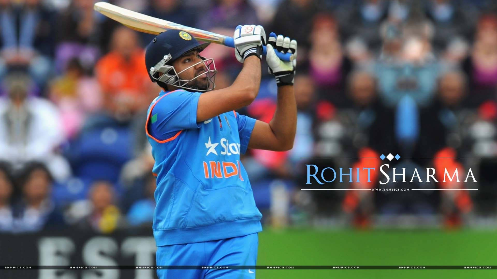 Rohit Sharma 桌布 壁紙 — 板球運動員 Rohit Sharma 身穿藍色印度球衣，在揮棒中途舉起球棒，背景是模糊的體育場人群。