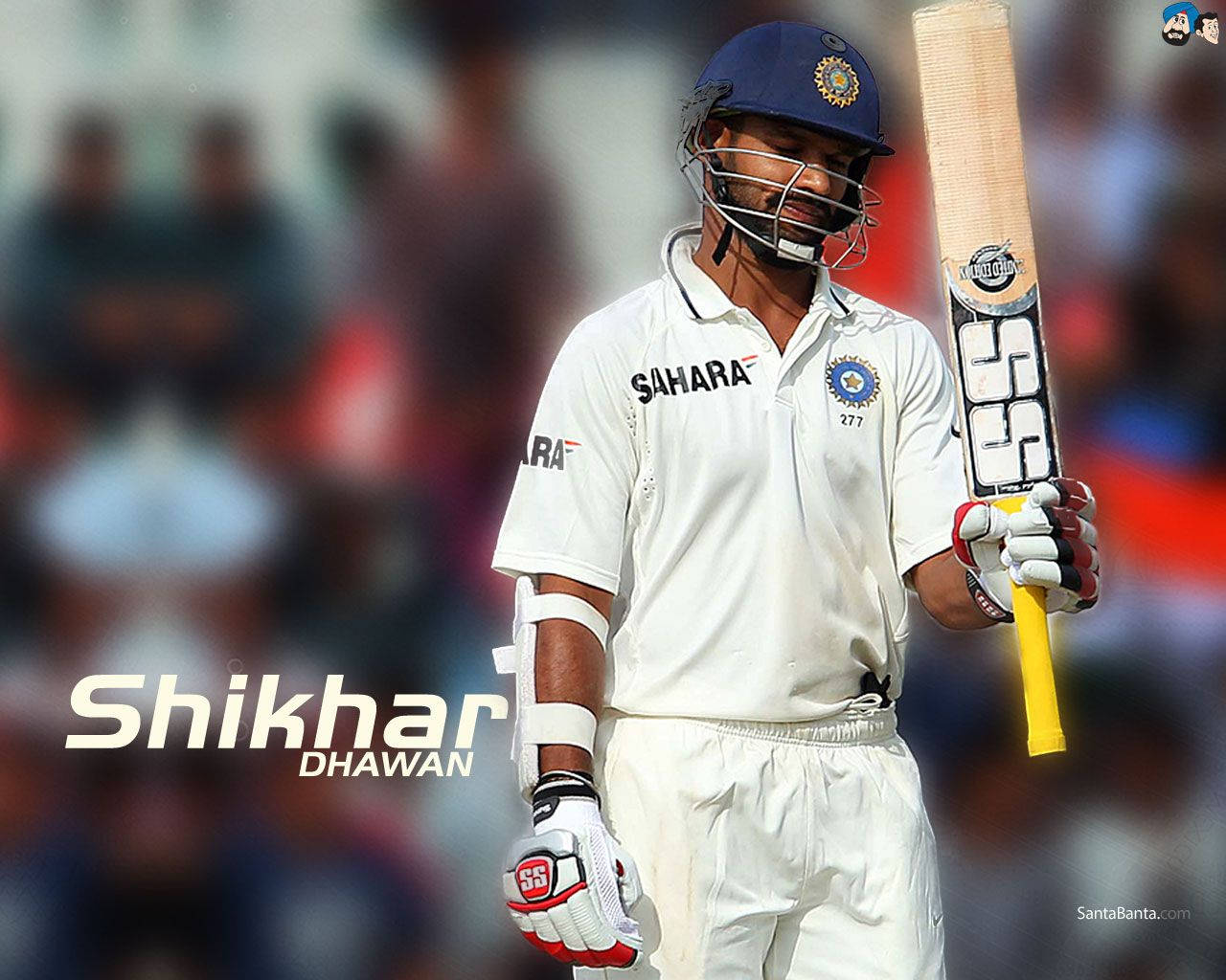 shikhar dhawan 桌布 壁紙 — 板球運動員 Shikhar Dhawan 身穿白色制服和藍色頭盔，手持球棒，表情專注，背景是模糊的體育場人群。