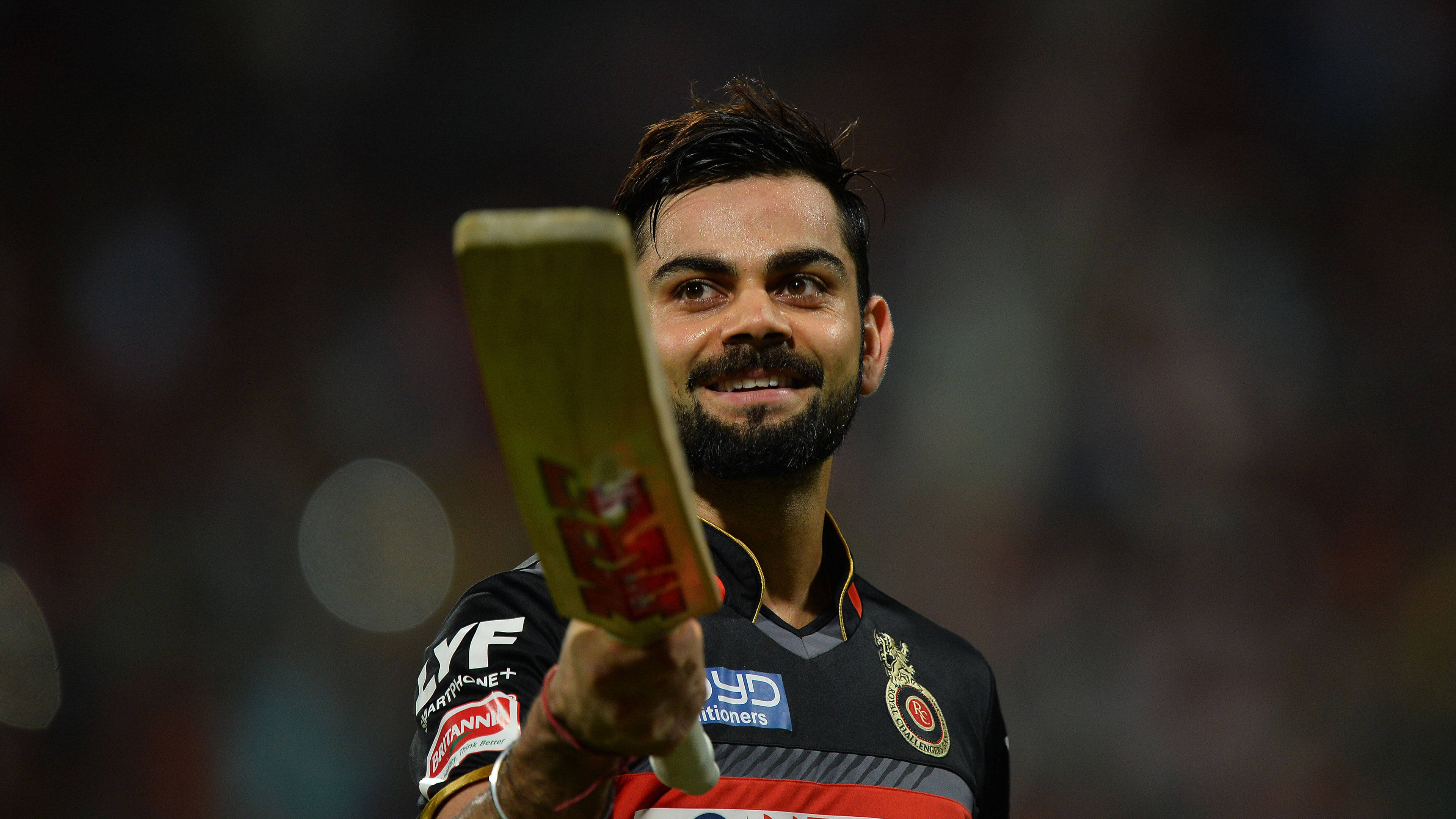 運動 桌布 壁紙 — 板球運動員 Virat Kohli 在夜間比賽中自豪地微笑，手持球棒，在體育場燈光下。