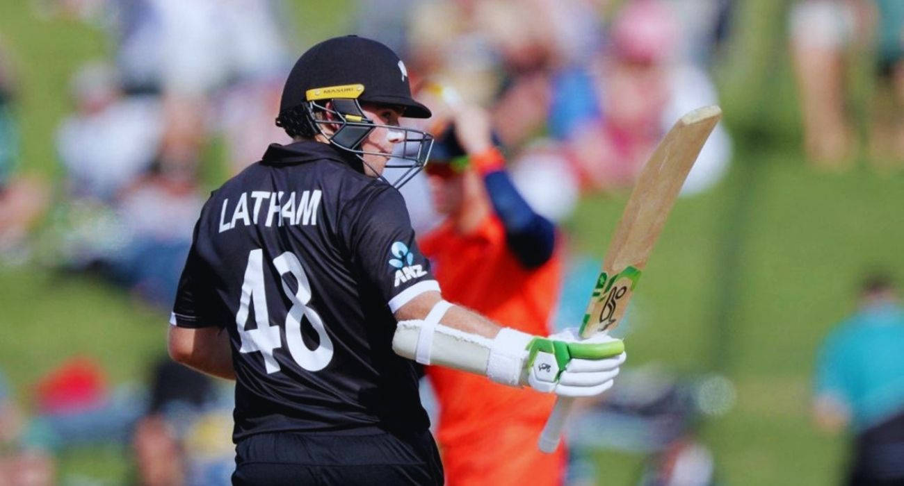 Tom Latham 桌布 壁紙 — 板球運動員Latham身穿黑色球衣48號，在陽光明媚的綠色球場上揮棒中，背景有模糊的觀眾。
