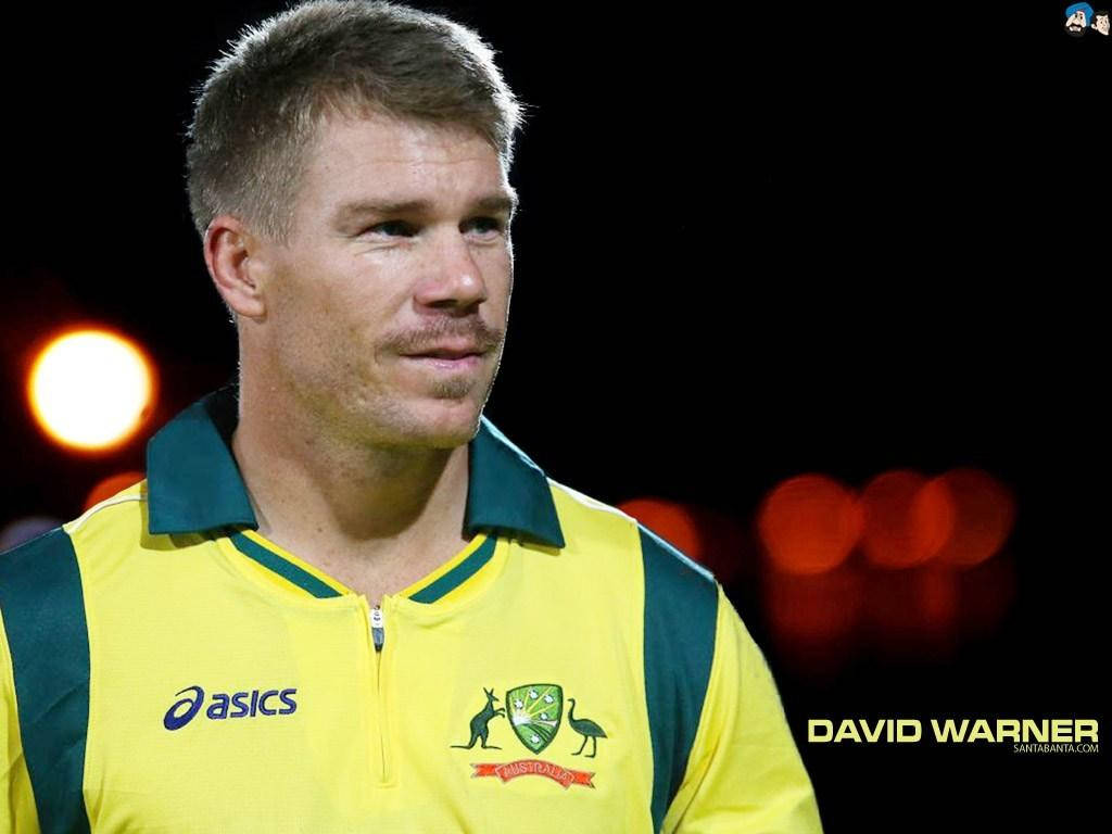 Warner 桌布 壁紙 — 板球運動員大衛·沃納的近距离肖像，身穿黃綠色澳洲球衣，背景是夜間模糊的體育場燈光。