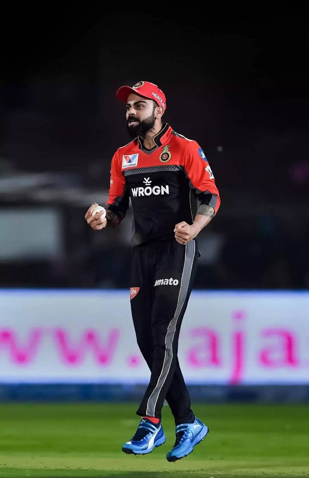 virat kohli rcb 桌布 壁紙 — 板球運動員維拉特·科利戴著紅色帽子和穿著紅色球衣，在體育場燈光下的綠色場地上邁步。