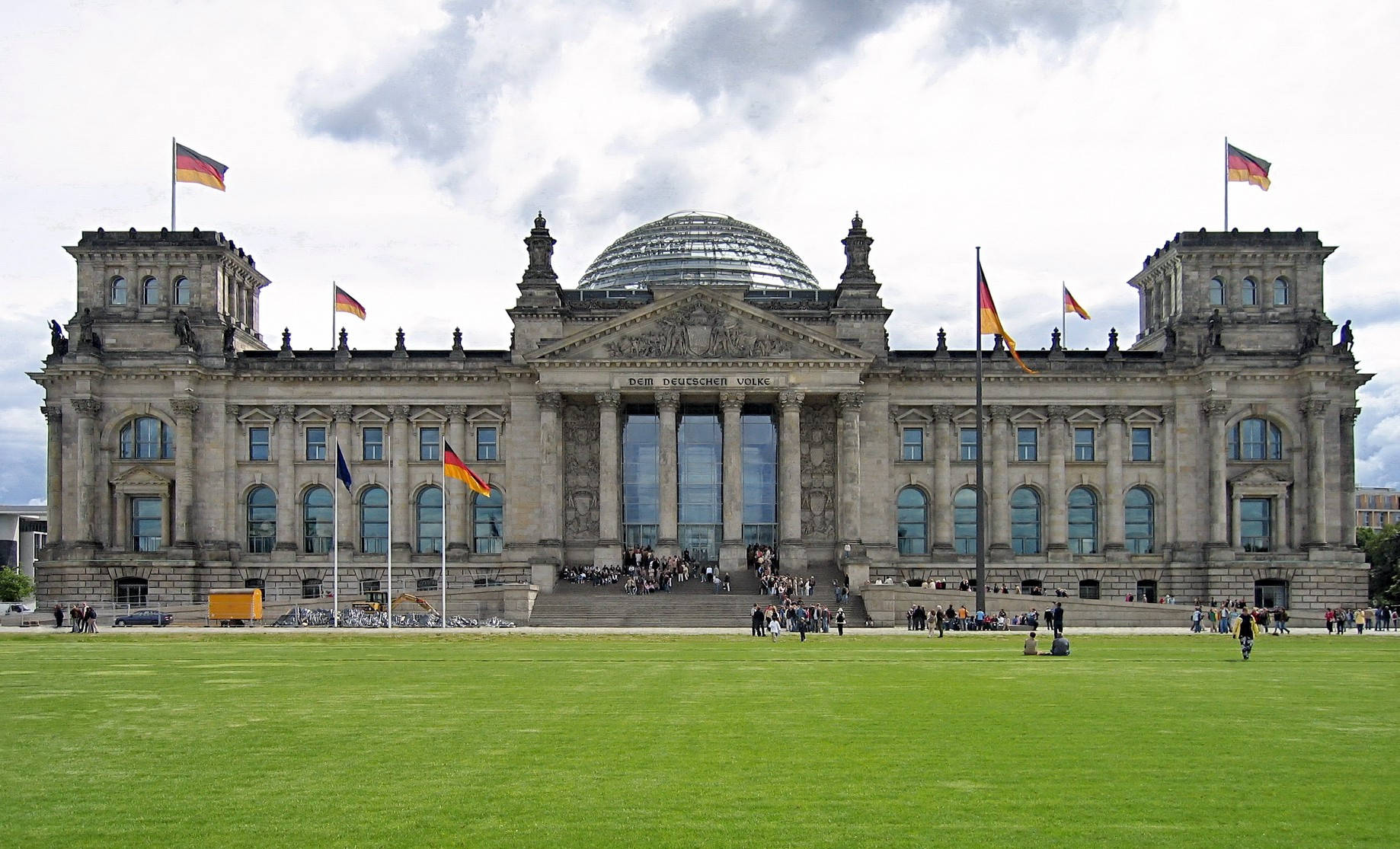 Berlin 桌布 壁紙 — 柏林國會大廈在陰天之下巍然聳立，玻璃圓頂閃閃發光，德國國旗在充滿活力的綠色草坪上空飄揚。