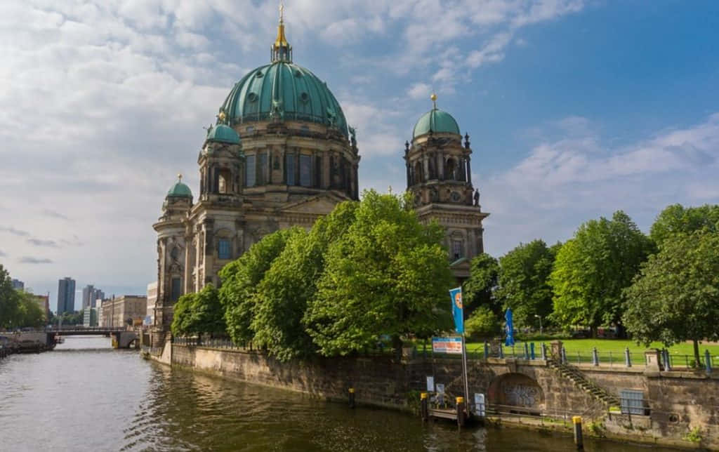 Berlin 桌布 壁紙 — 柏林大教堂的綠色圓頂在部分多雲的天空下，雄偉地矗立在兩旁綠樹成蔭的河邊。