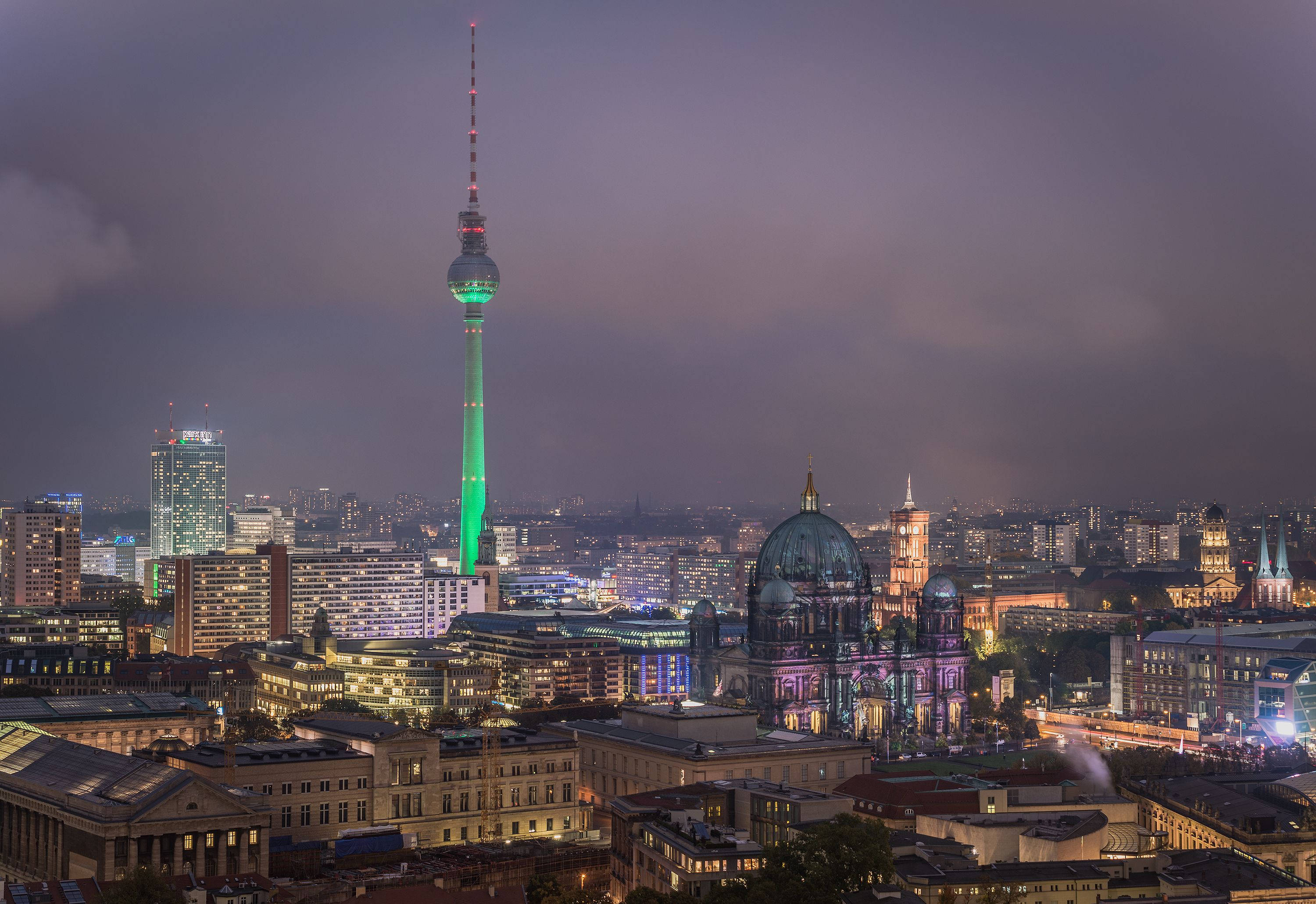 Berlin 桌布 壁紙 — 柏林天際線在黃昏時分，標誌性的電視塔發出綠色光芒，圓頂大教堂在陰鬱的紫色天空下被照亮。